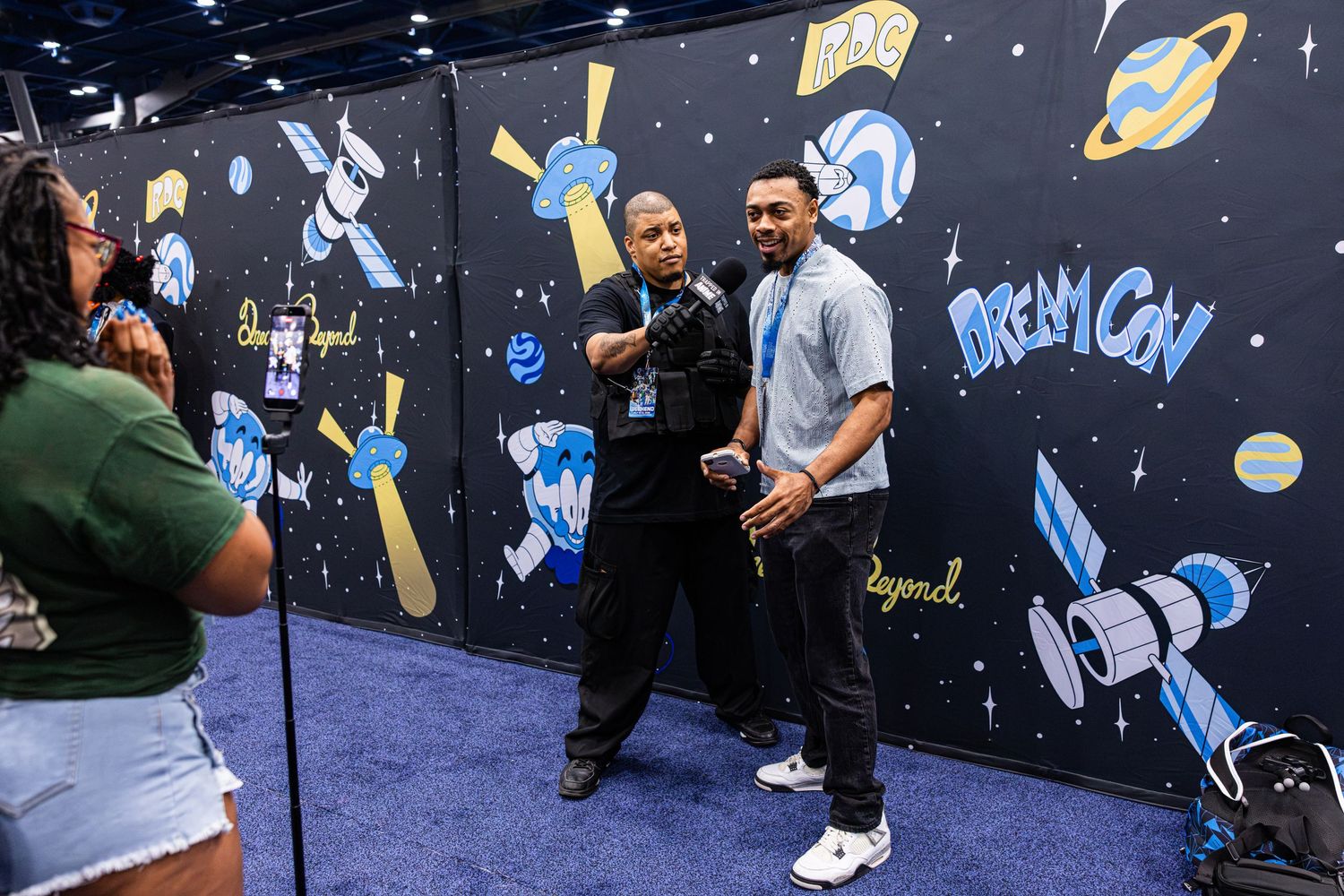 Two men pose for a photo at DreamCon event in front of a colorful space-themed mural with rockets and planets.