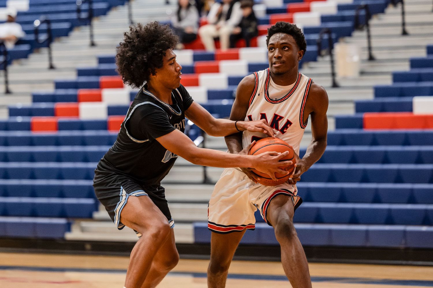 Two basketball players compete for possession during a high school game with red and blue bleachers in the background.
