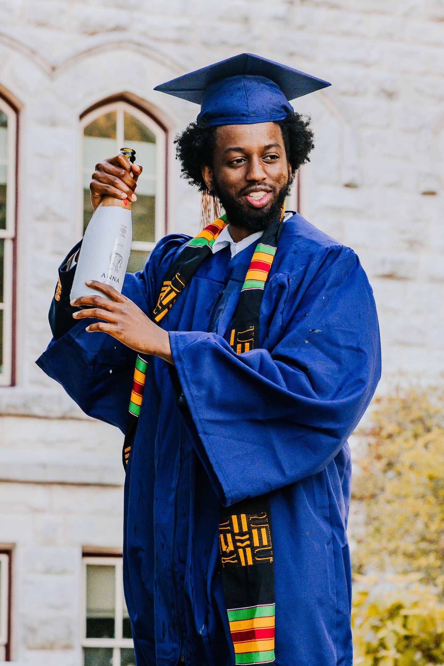 Graduate in blue cap and gown with colorful kente stole celebrates holding diploma on sunny campus day.