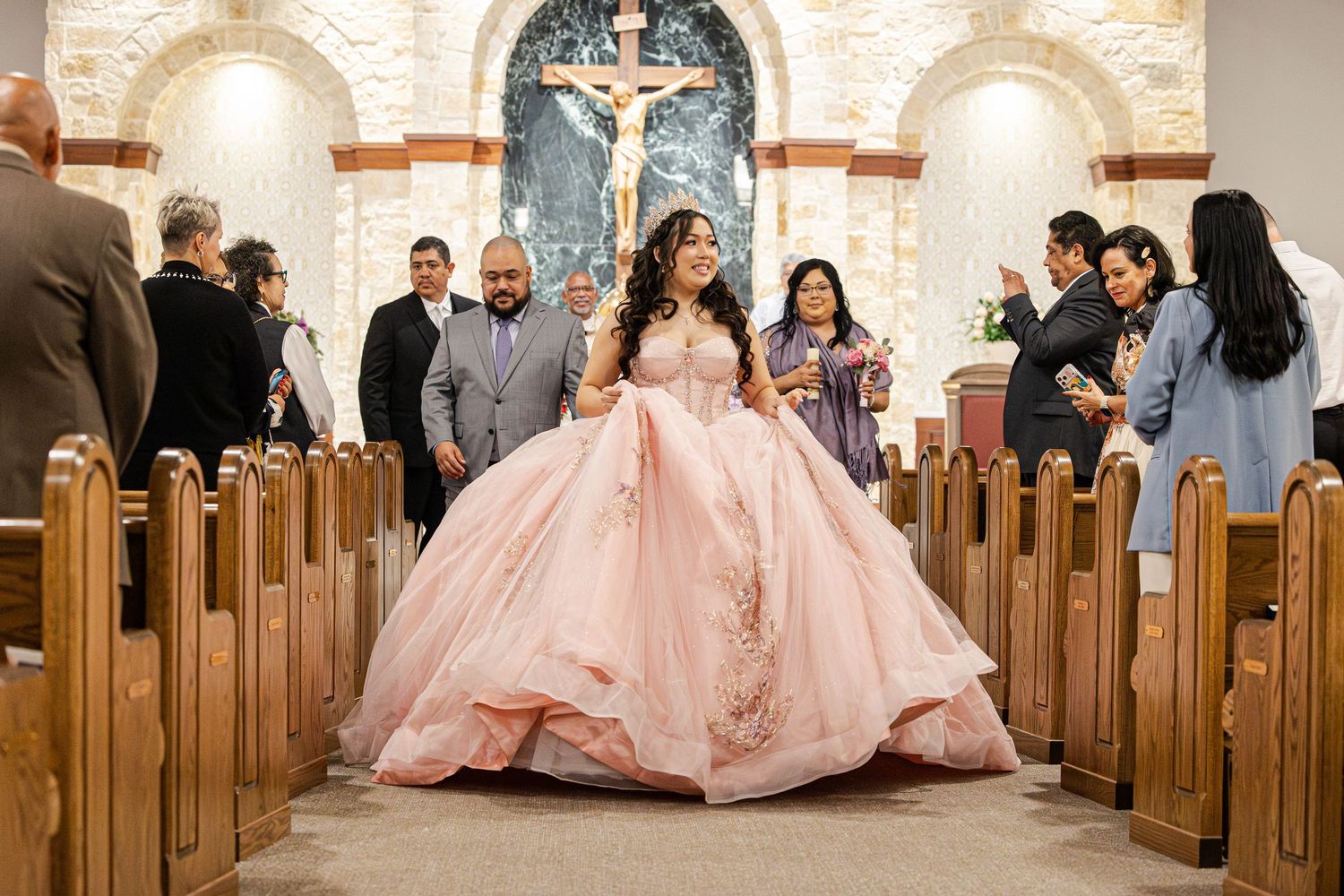 Wedding party members in pink tulle dresses gather in wooden church pews during a formal ceremony.