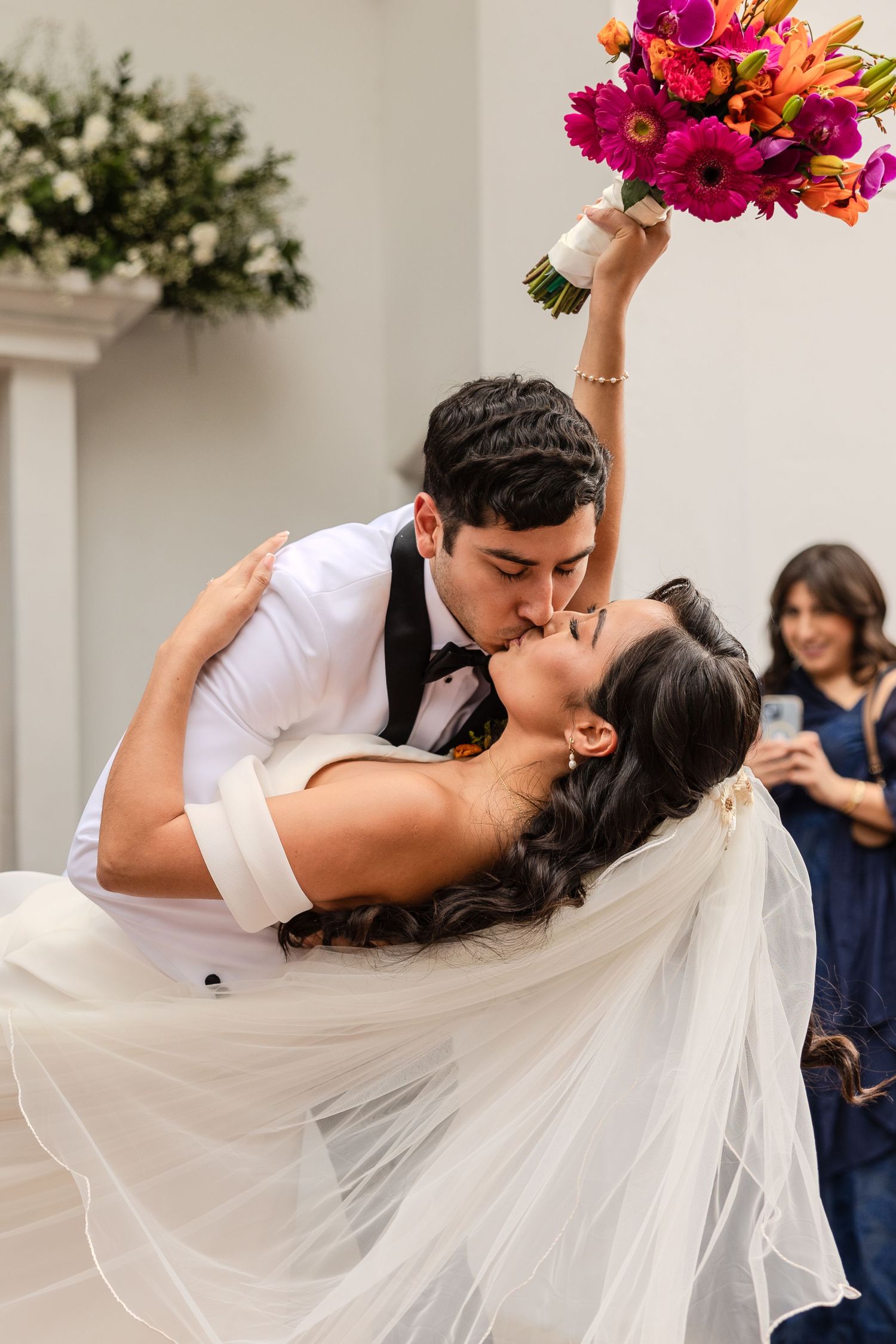 Newlyweds share a romantic dip kiss with vibrant pink and orange bouquet during their wedding celebration.