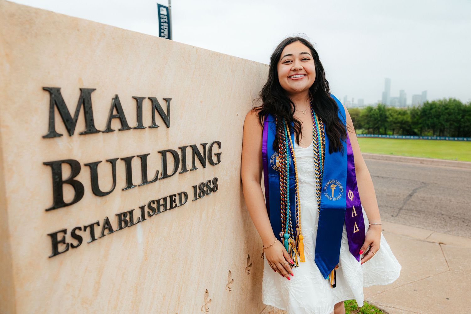 A smiling graduate in cap and gown poses beside the Main Building Established 1888 sign on a university campus.