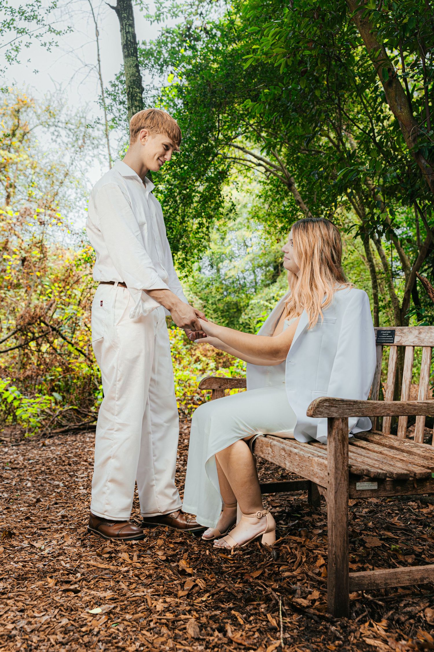 A romantic outdoor engagement photo series showing a couple sitting on a rustic wooden bench surrounded by trees and leaves.