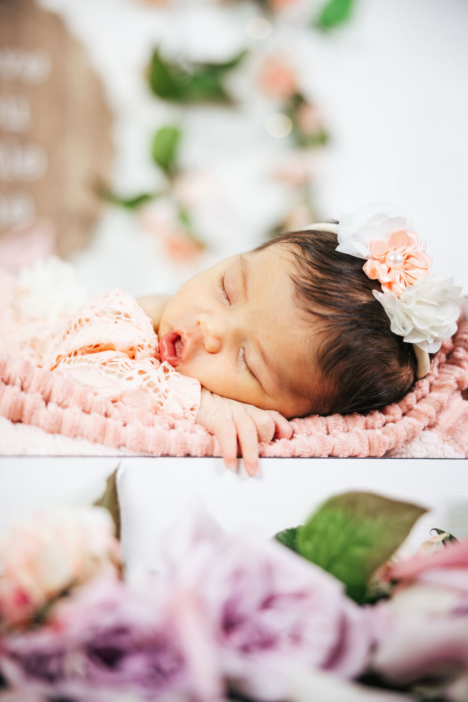 Sleeping newborn baby wrapped in pink blanket with delicate floral decorations and greenery in soft lighting.
