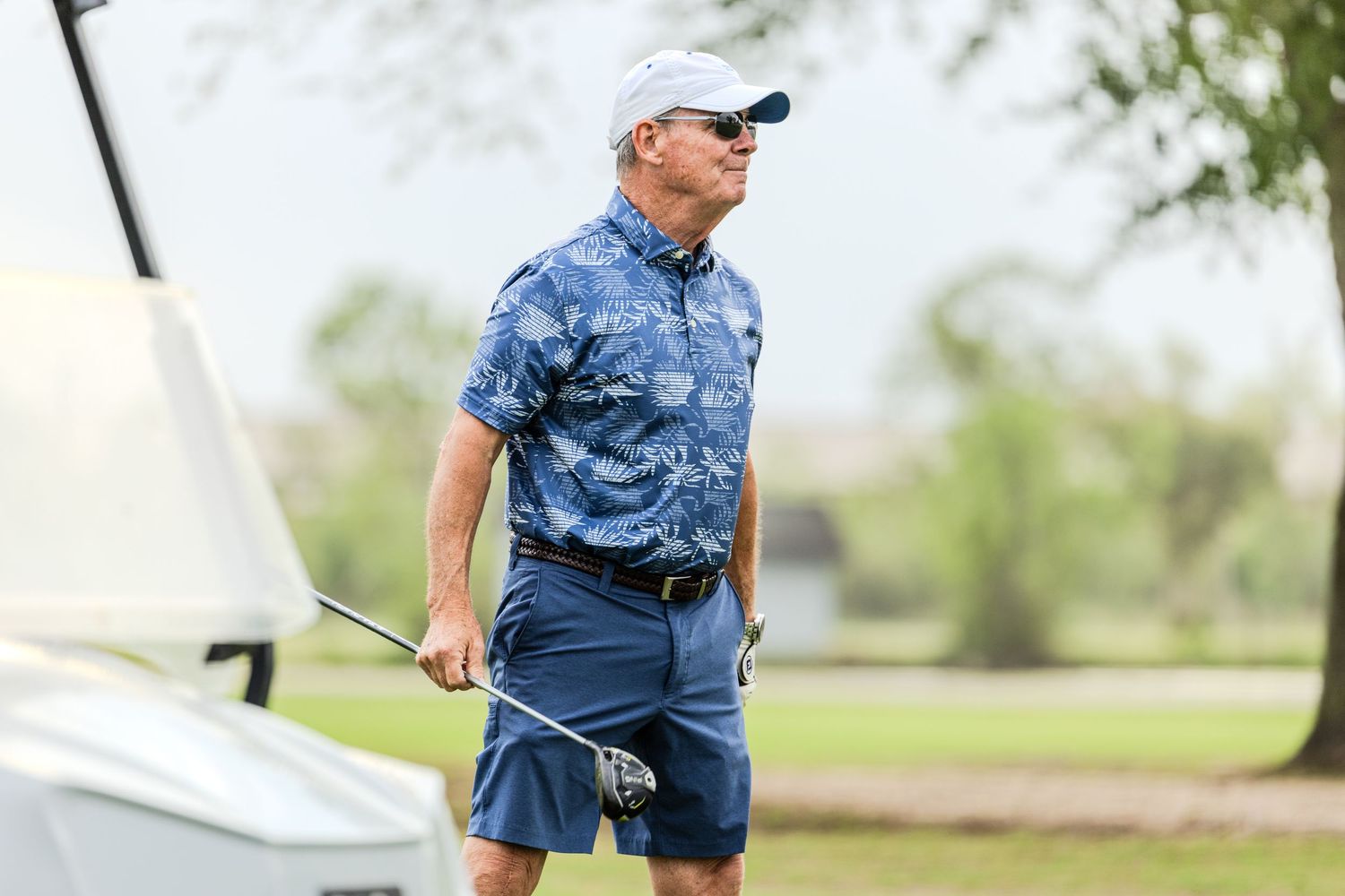 A golfer in a blue polo shirt takes a swing on a sunny golf course with trees in the background.