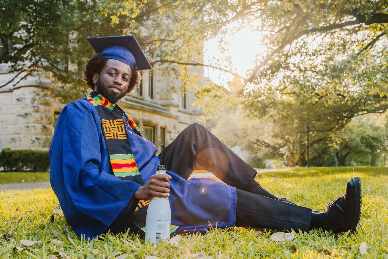 A person in a blue hoodie relaxes on autumn grass while reading and enjoying a beverage outdoors.