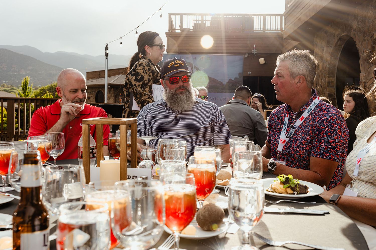 People dining outdoors at a long table with drinks, enjoying conversation in a scenic mountain setting.