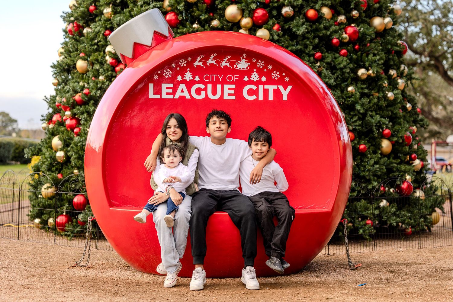 Family portrait session in front of a large red Christmas ornament prop with a decorated tree in the background.