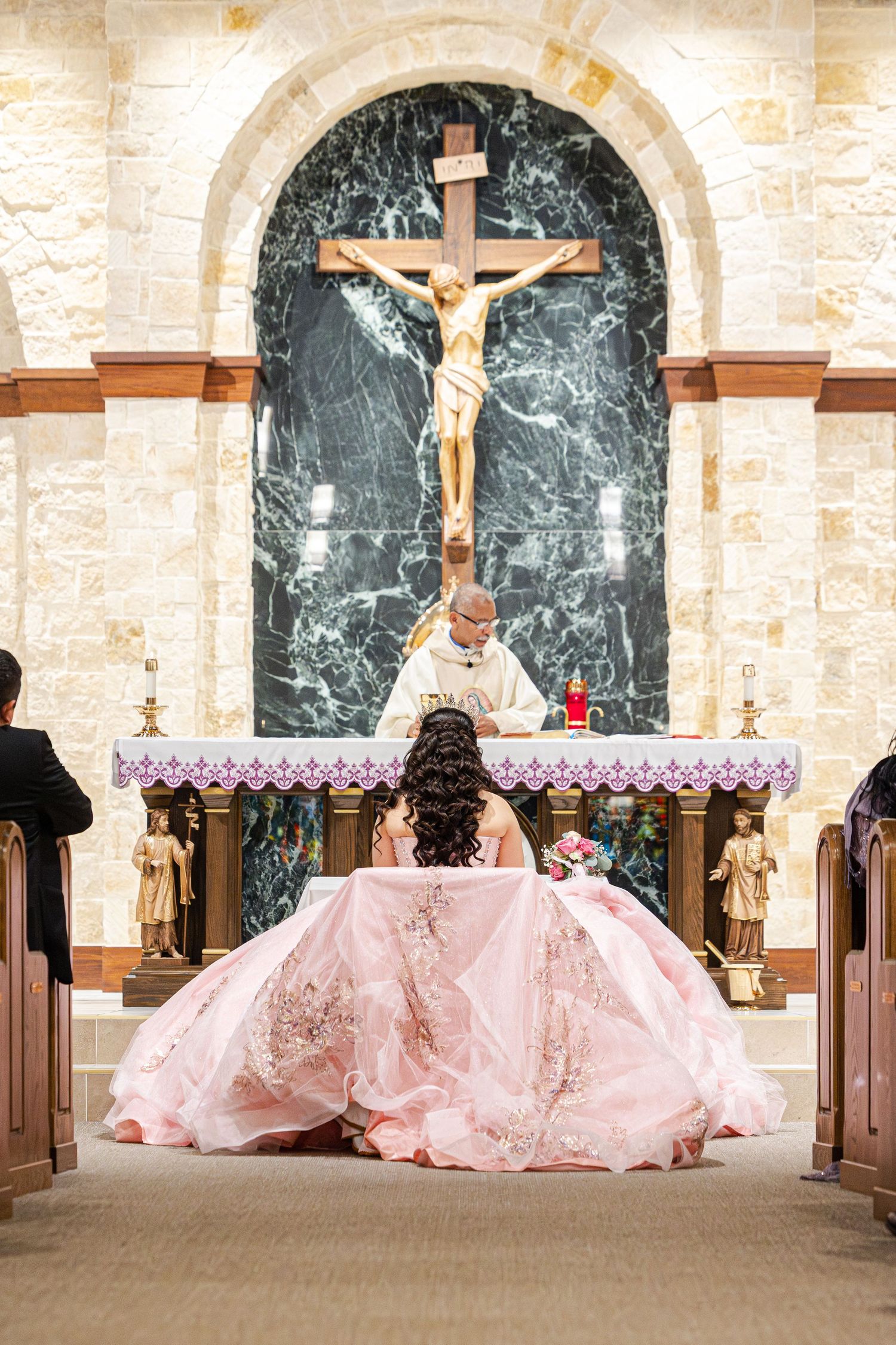 Pink ballgowns create a graceful scene as people bow in prayer at a church altar with marble columns and crucifixes.