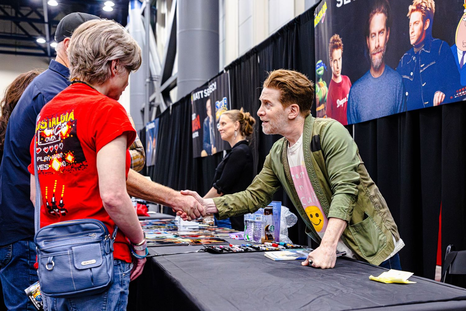 Celebrity signing autographs and shaking hands with fans at a convention table indoors.