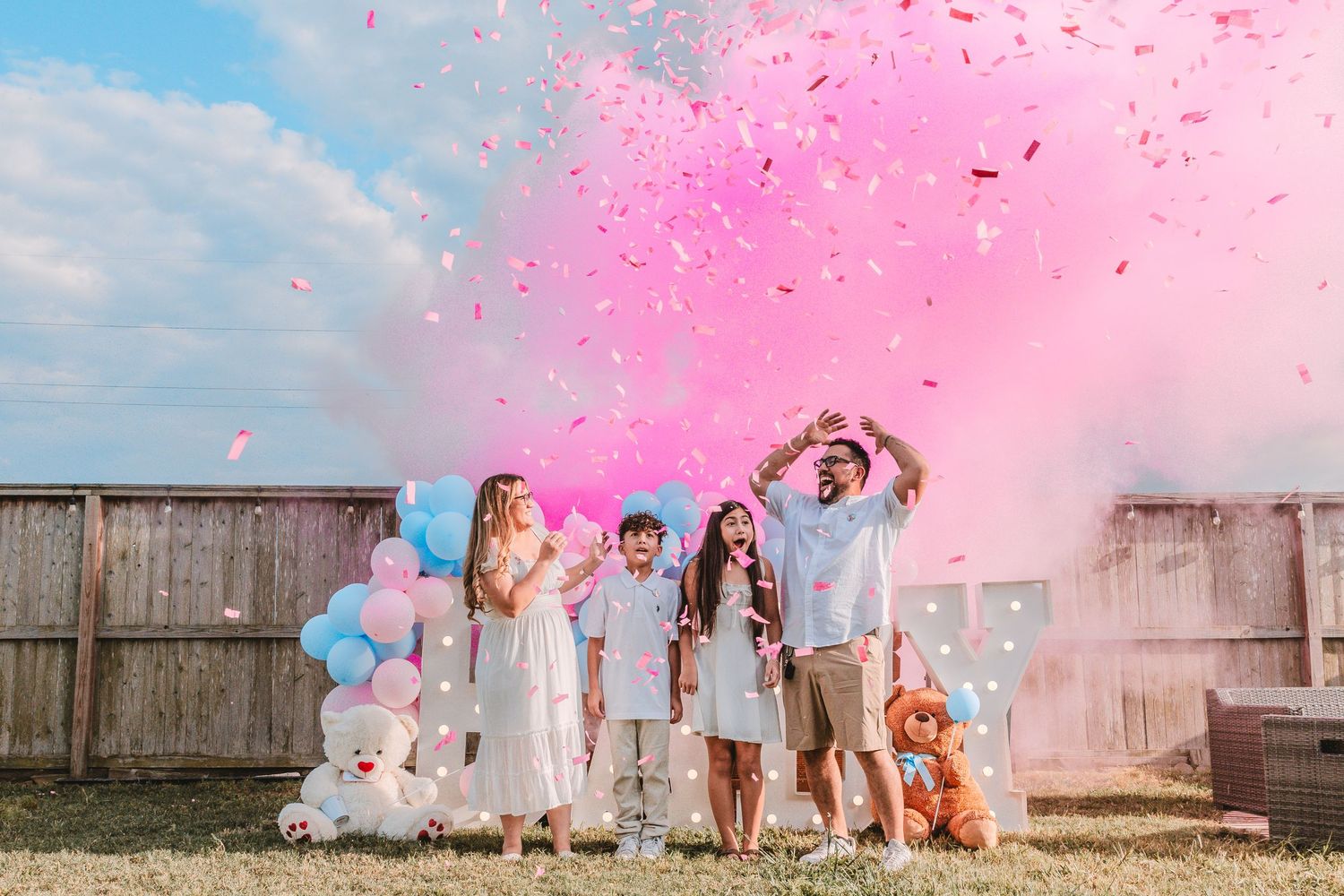Family gender reveal party with pink confetti cannon, balloons, and excited children in a backyard.