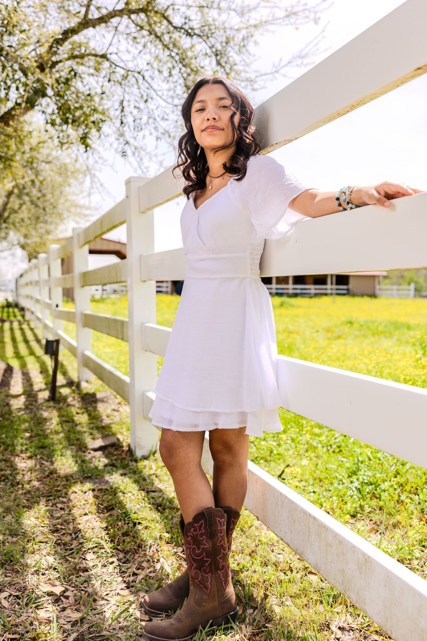 A person in a white dress and brown cowboy boots poses playfully along a white fence in a sunny field.