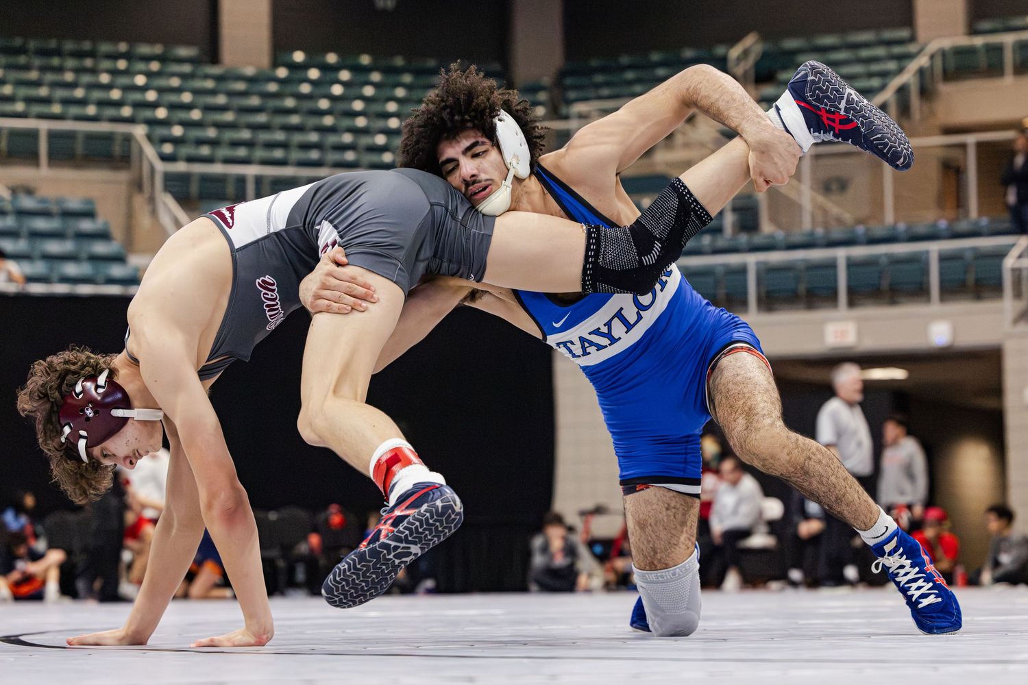 Two wrestlers compete in a high school or collegiate wrestling match on a mat with spectator seating visible in background.