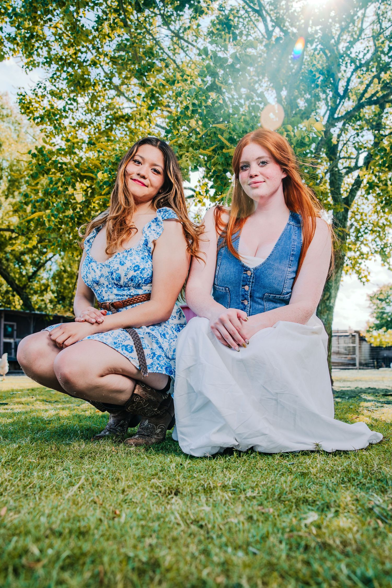 Two friends in blue and white dresses sit together on sunny grass during a summer photoshoot.