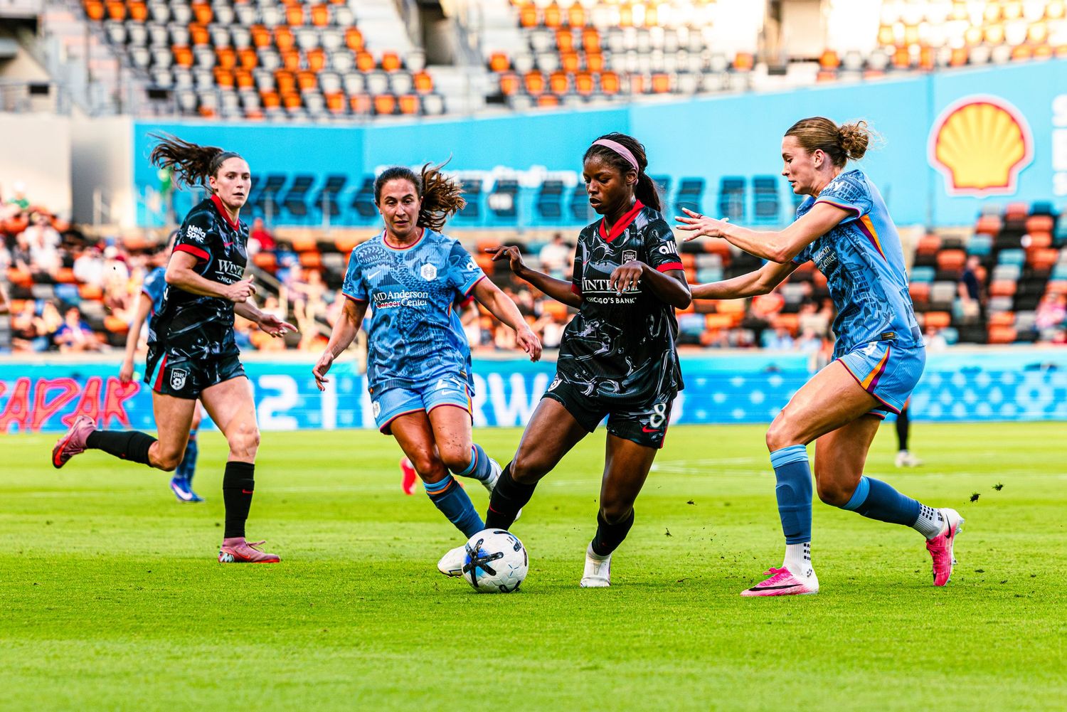 Women soccer players compete for the ball during an intense match on a bright green field with orange stadium seats.