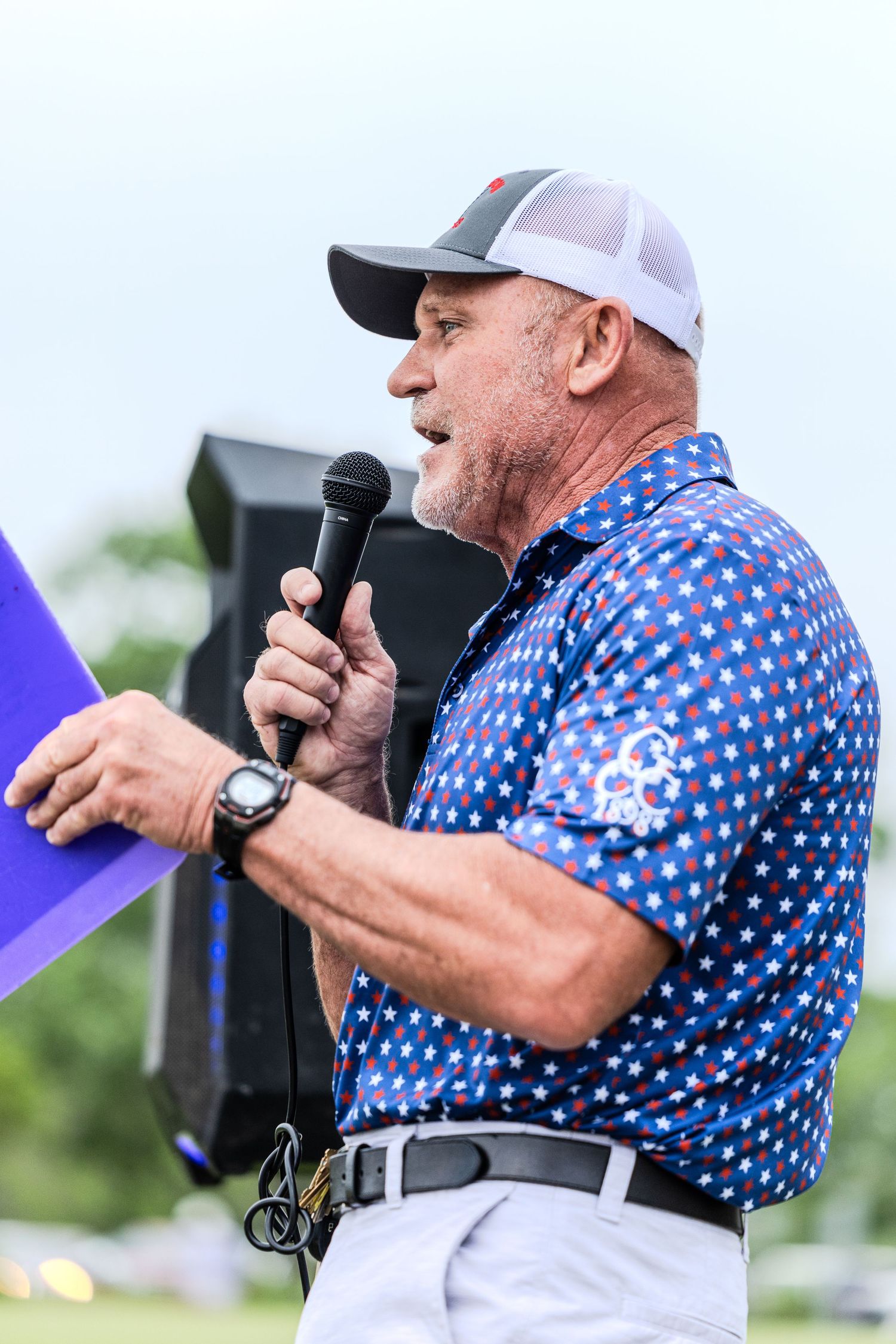 Performer in a blue polka dot shirt and white baseball cap sings into a microphone at an outdoor event.
