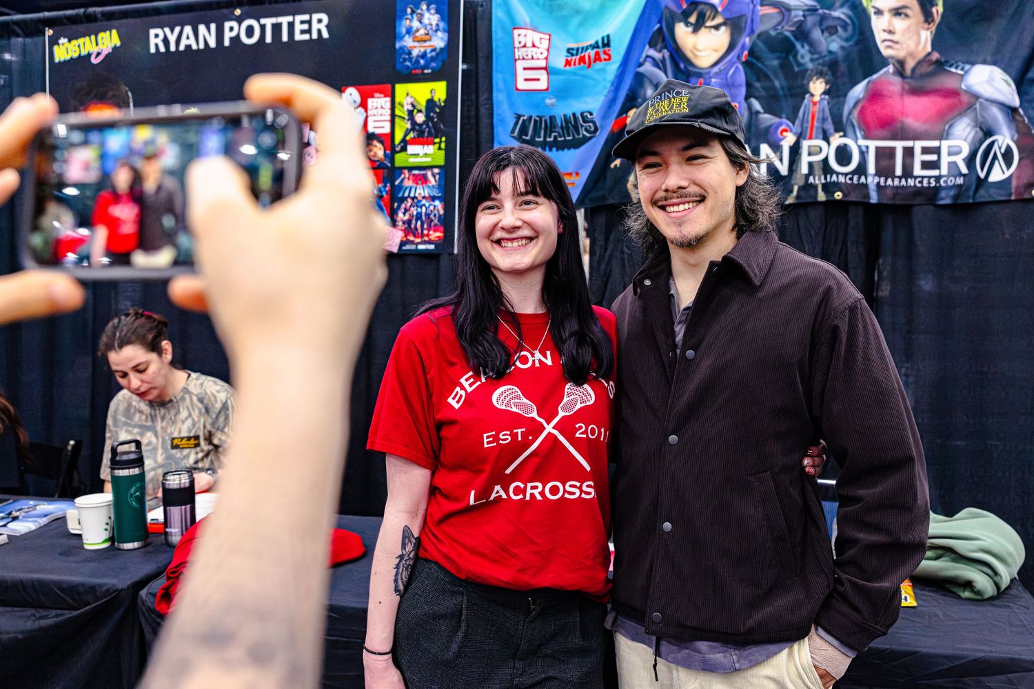 Fan in red lacrosse shirt poses for photo with Ryan Potter at comic convention booth.