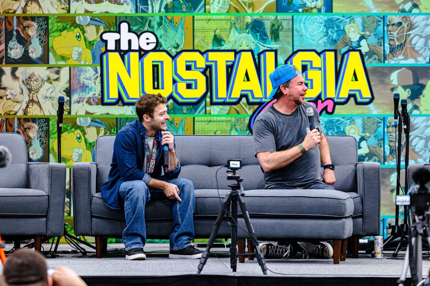 Two men talk on stage at The Nostalgia Show, seated on a couch with microphones against a colorful backdrop.