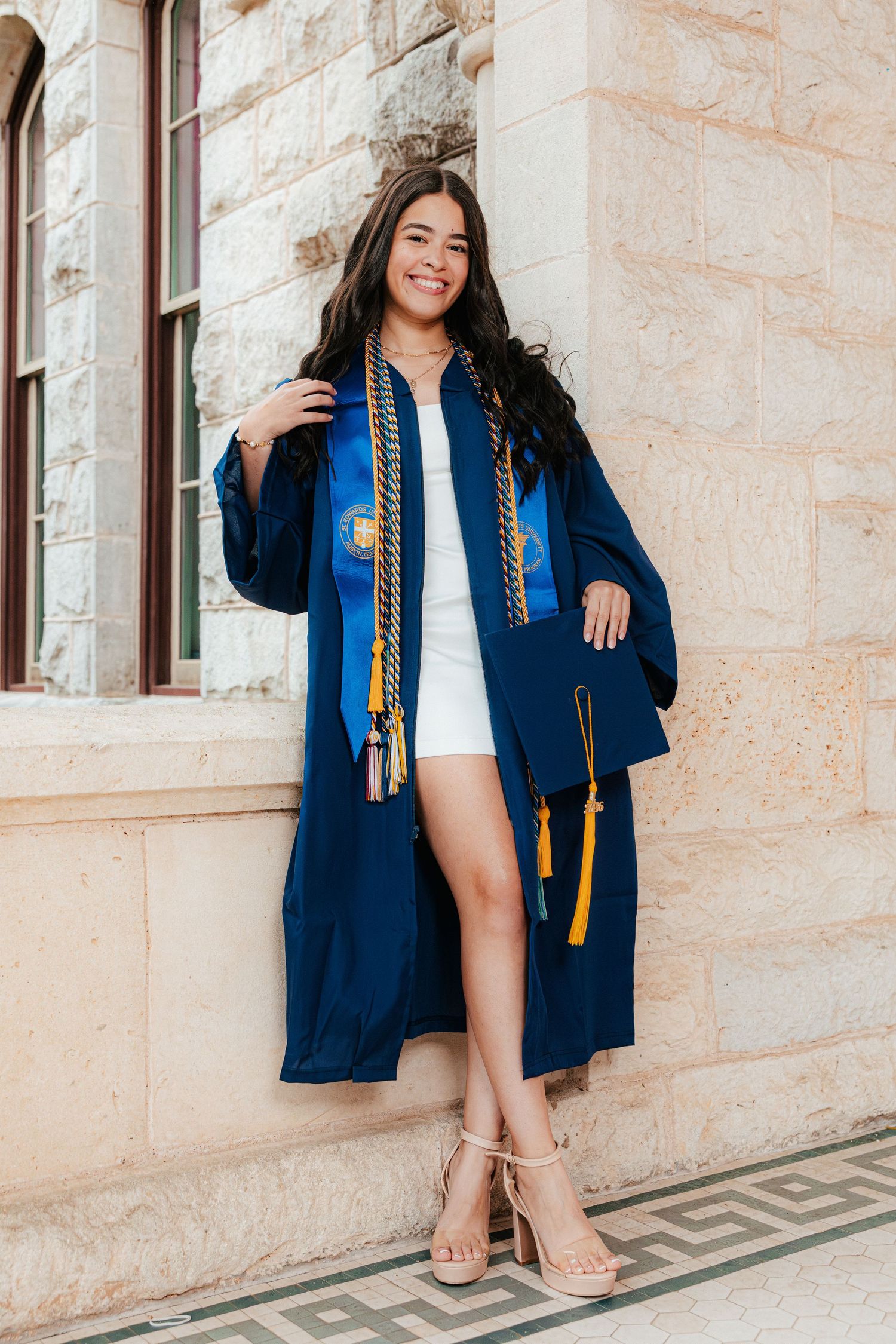 Smiling graduate in blue cap and gown with honor cords poses against stone building in white dress and heels.