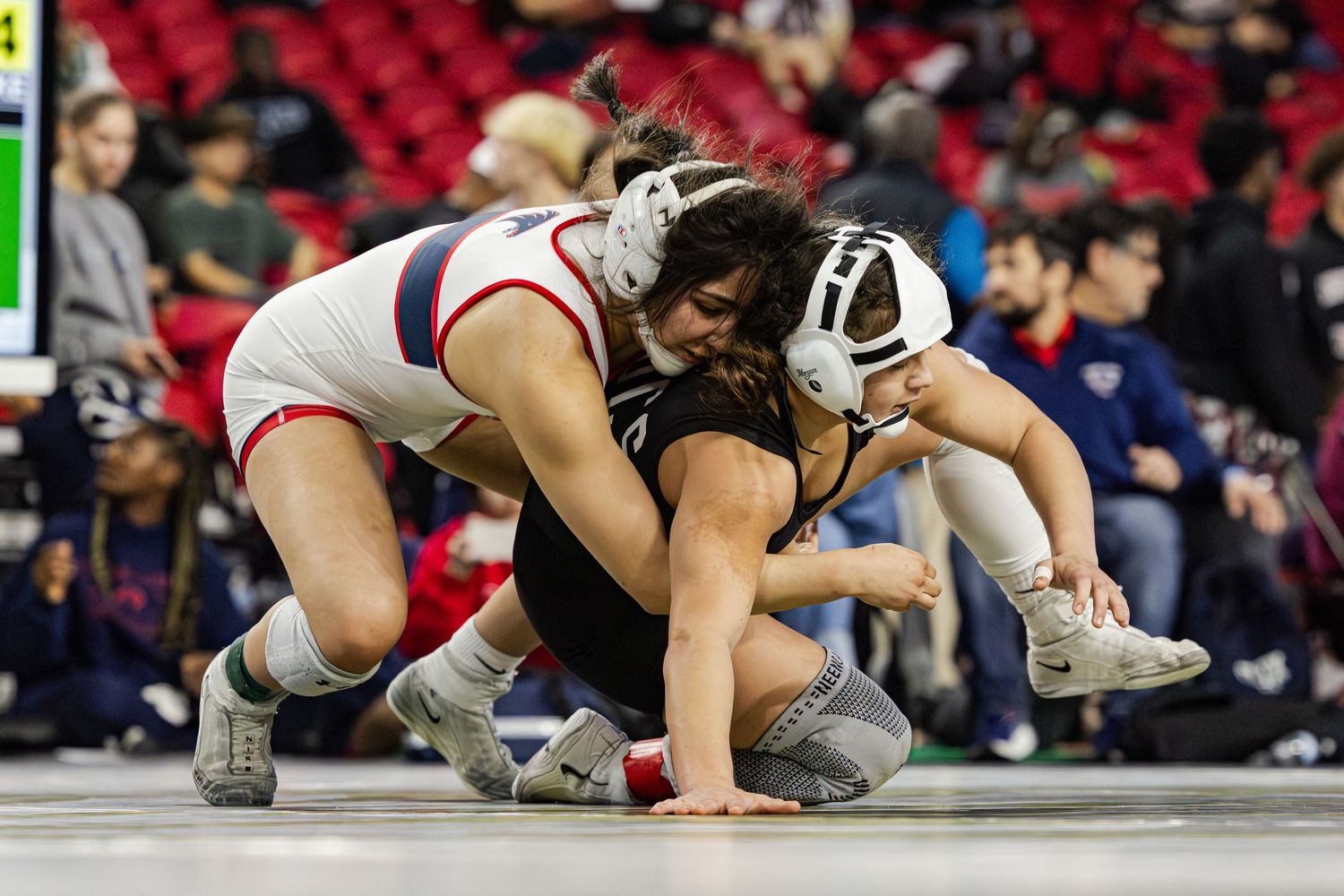 Two female wrestlers compete intensely on a mat during a wrestling tournament, one in white uniform grappling the other in black.