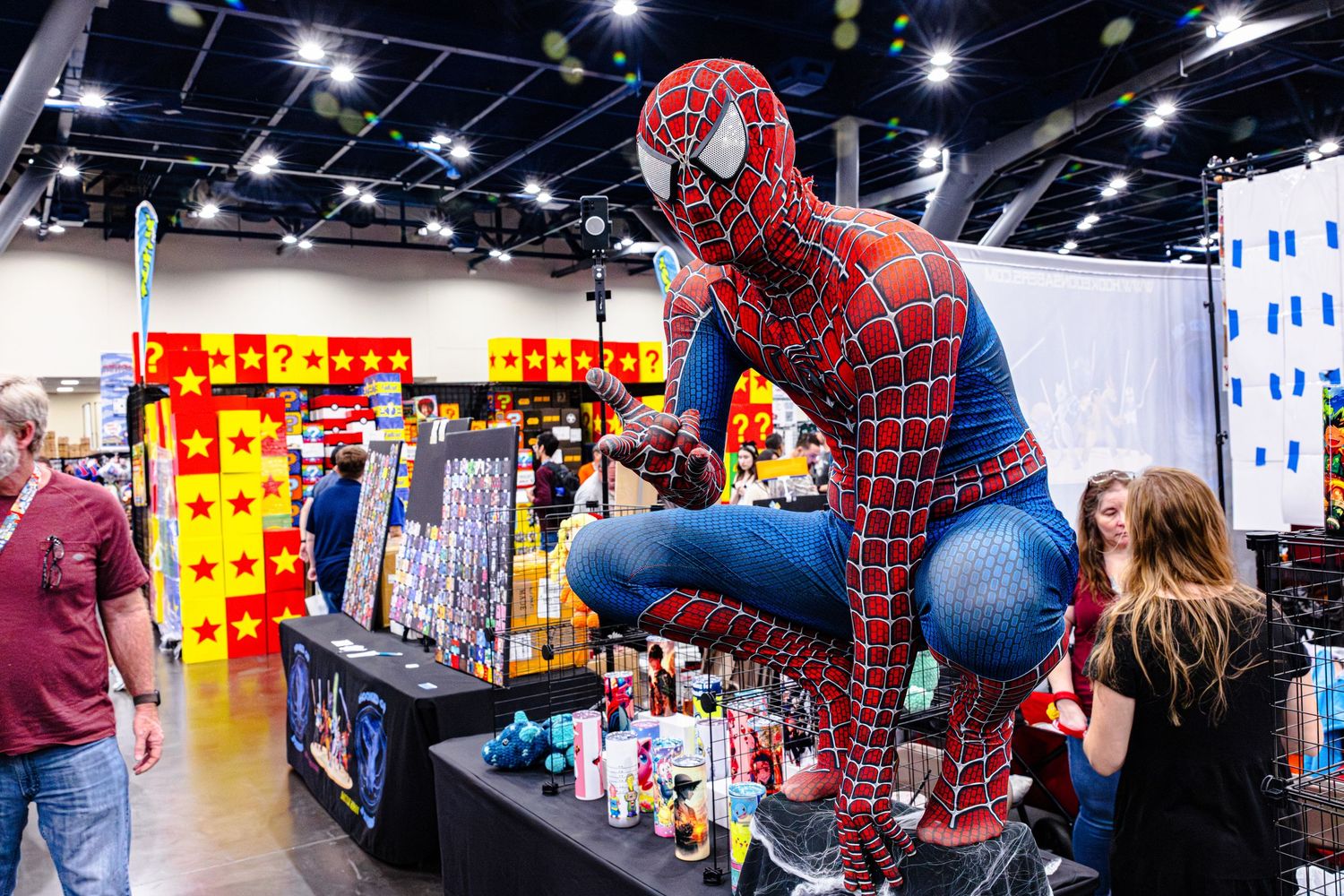 Person in Spider-Man costume crouches on display table at a comic book convention.