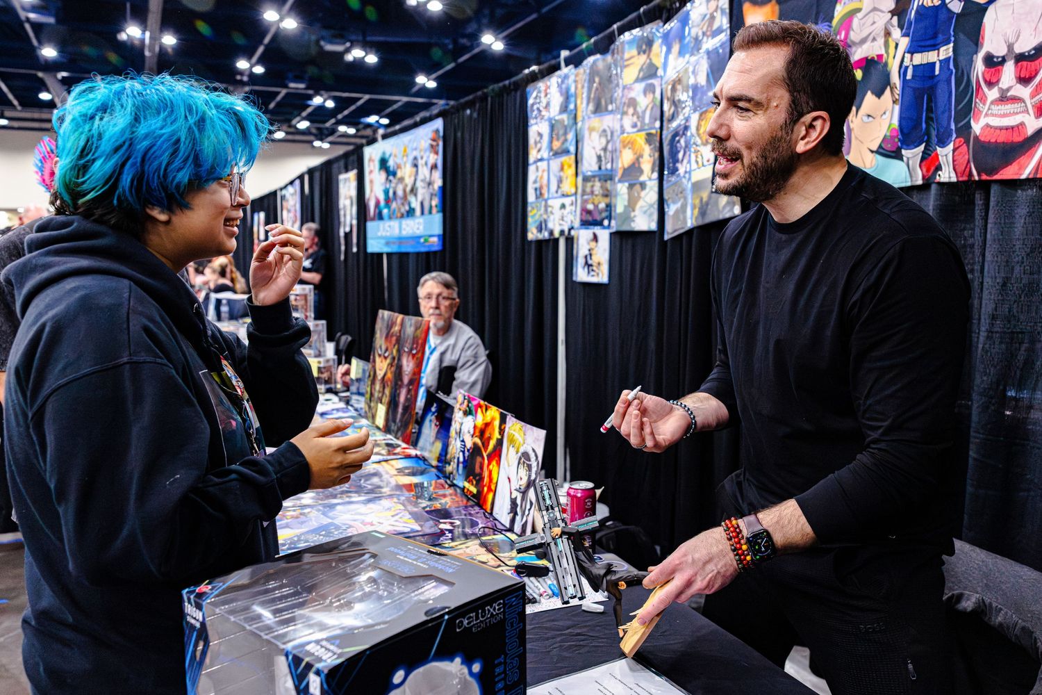 Man in black shirt engages in lively conversation with blue-haired attendee at comic con booth.