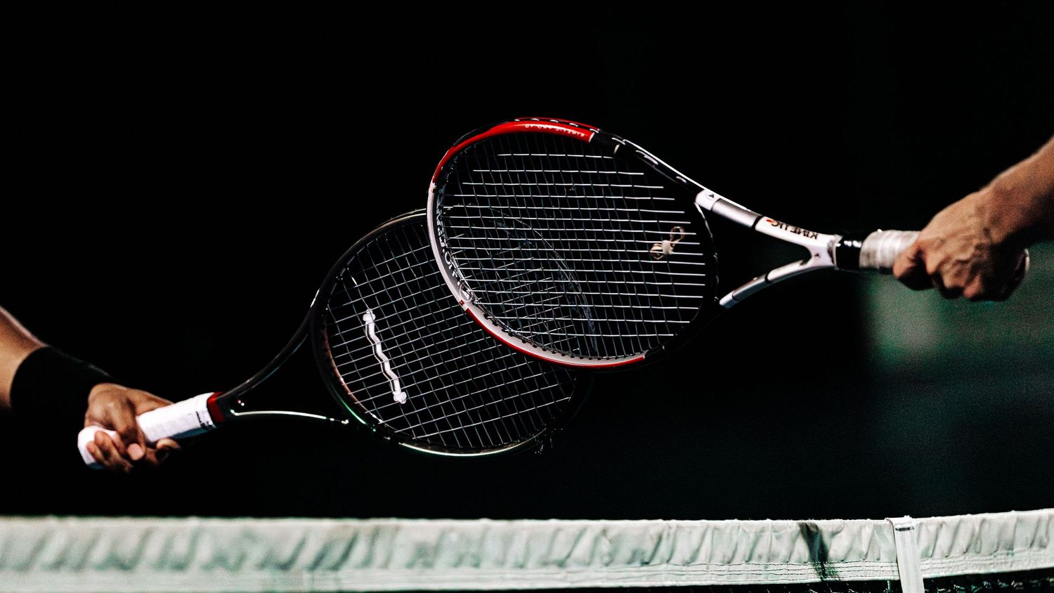 Two tennis rackets held by hands clash together in a dramatic action shot against a dark background.
