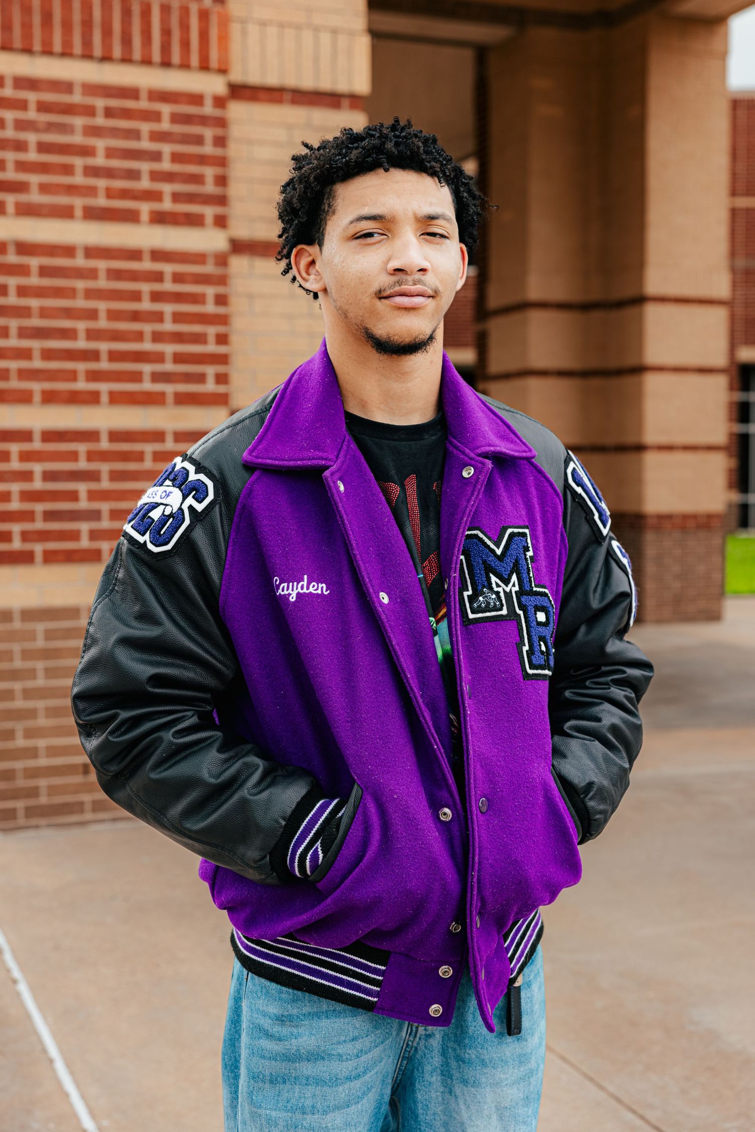 Young man wearing a purple varsity letterman jacket with black leather sleeves and name Kayden embroidered, standing outdoors.
