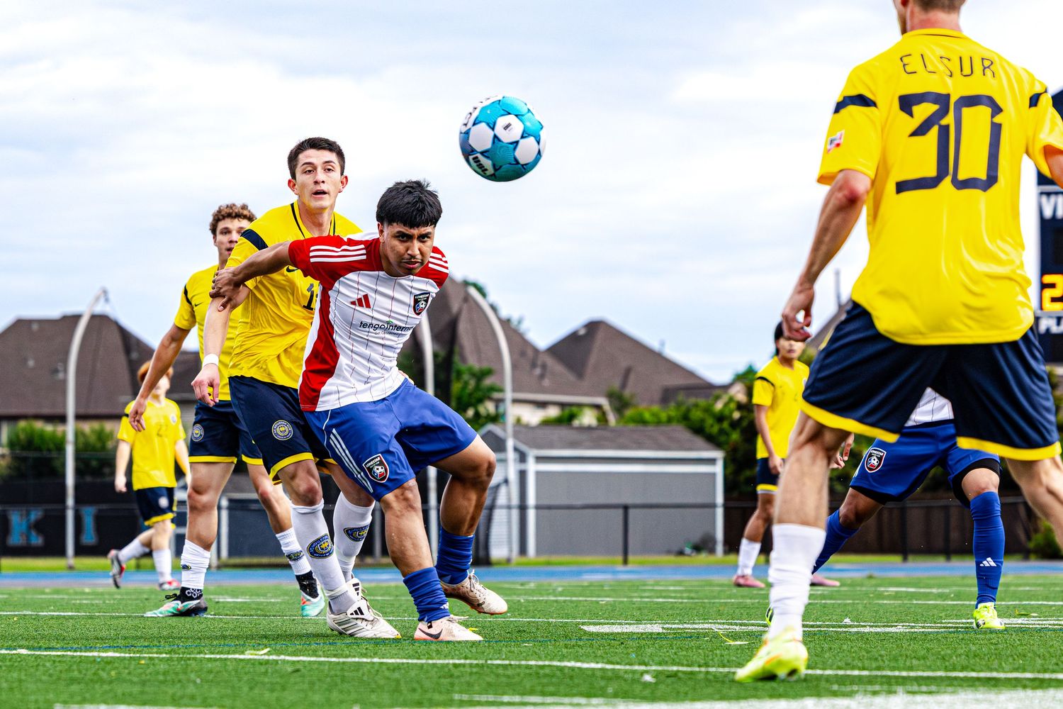 Soccer players compete for a header during a match, with players in blue and yellow uniforms on a turf field.