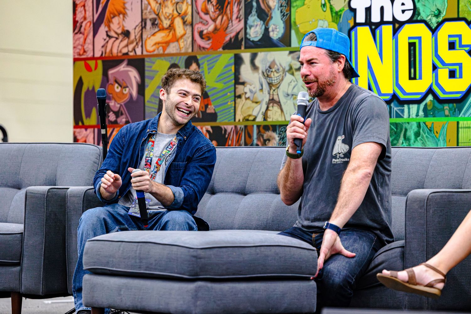 Two men laughing on a gray couch at a comic convention panel, one holding a microphone.