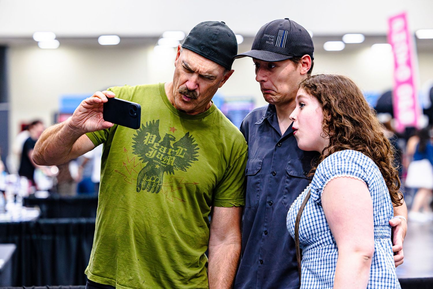 Two men and a woman examine a smartphone screen together at an indoor convention event.