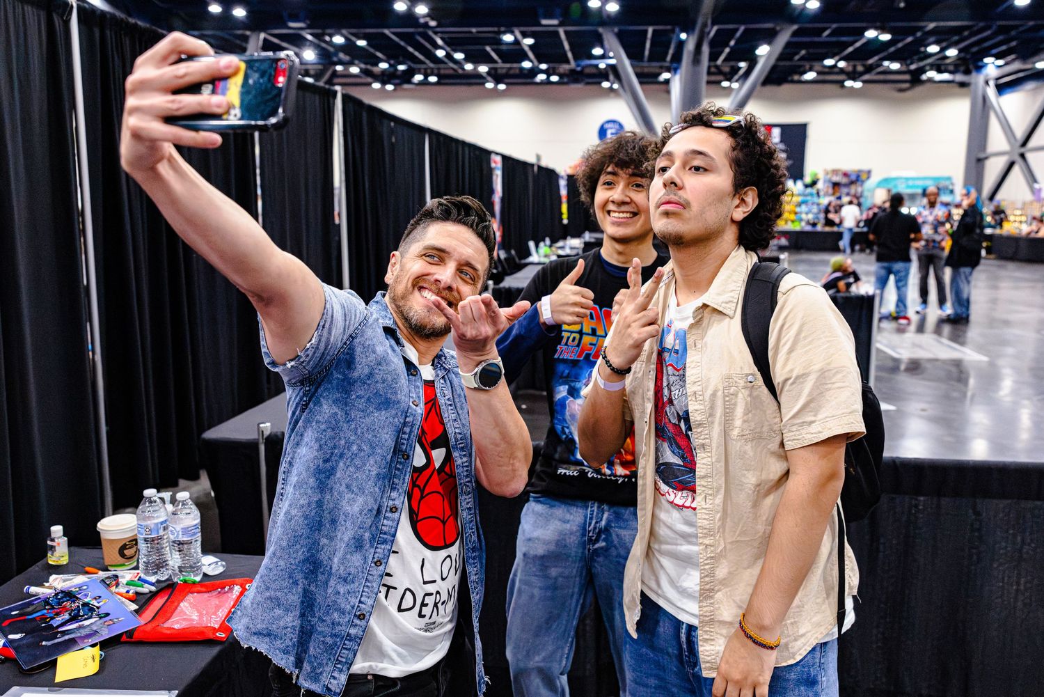 Three friends take a selfie together at a comic convention, flashing peace signs and smiling.