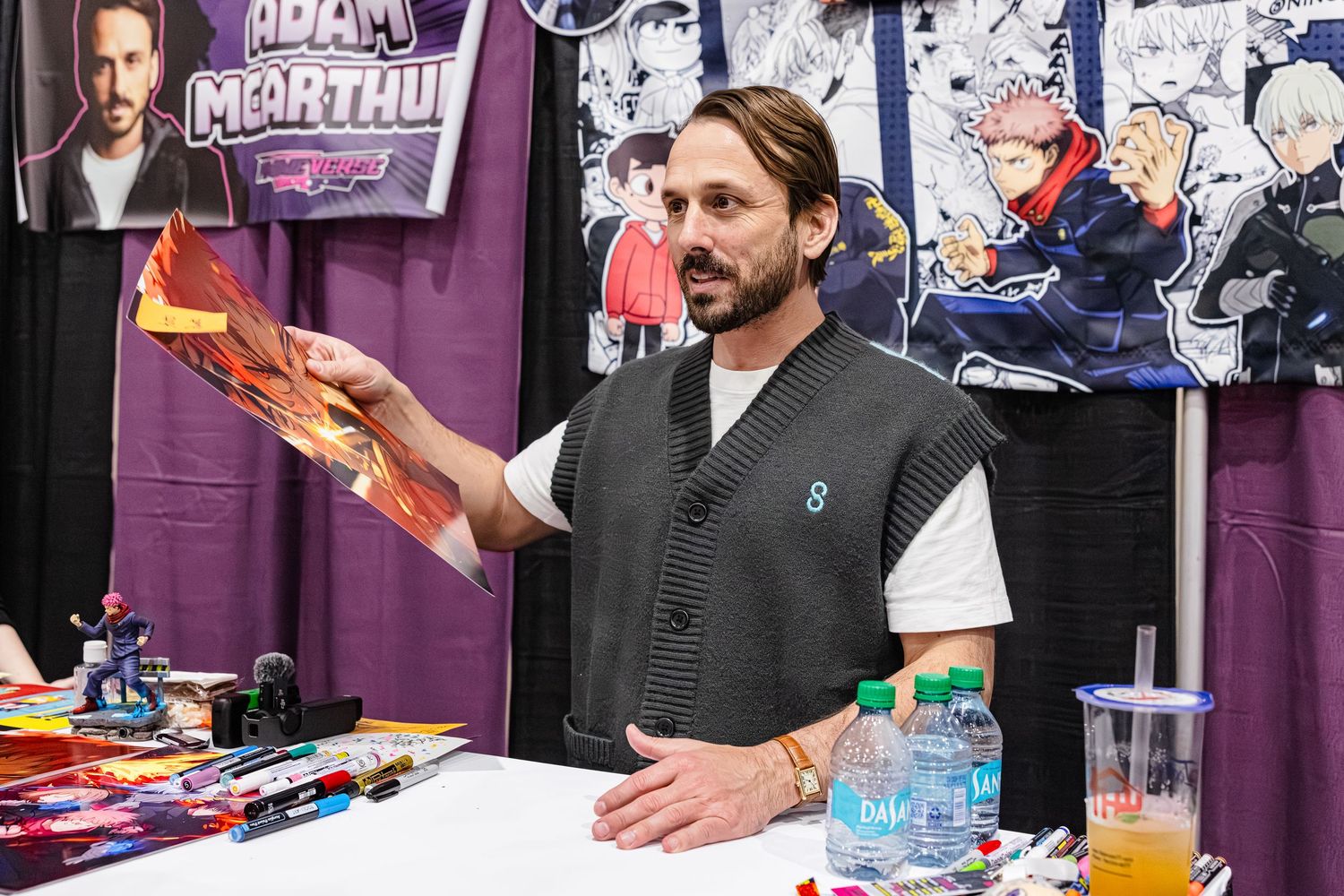 Man in gray vest holds artwork at comic convention booth with anime banners displayed behind him.