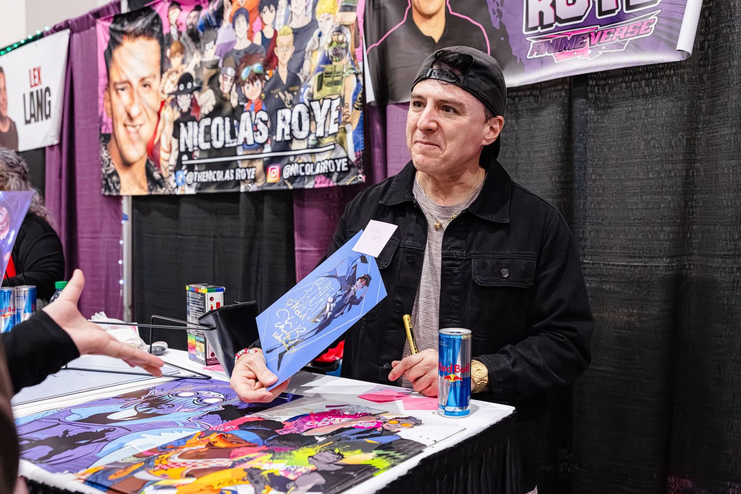 Nicolas Royz signing autographs at a convention booth with colorful artwork and Red Bull cans on display.
