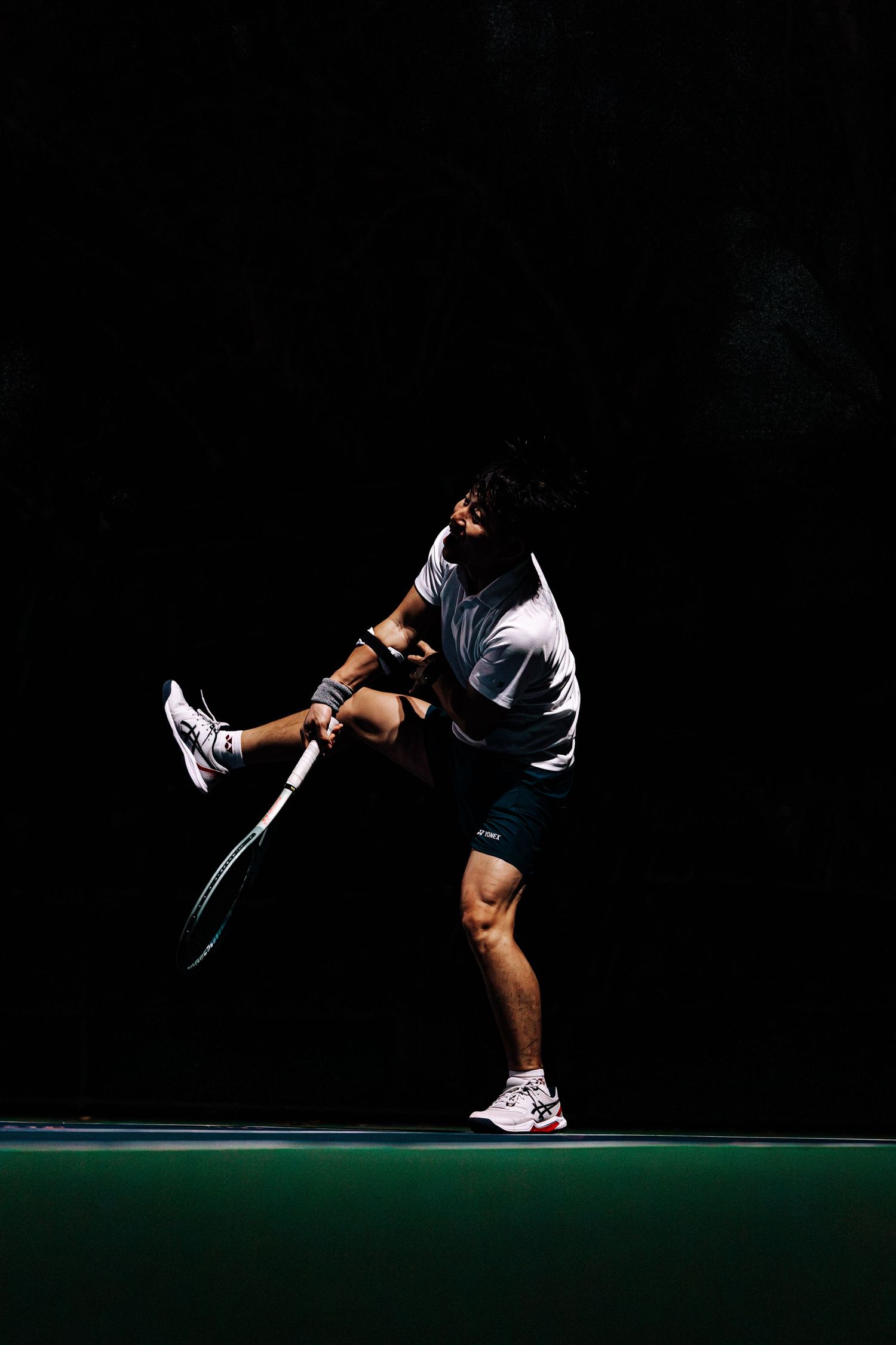 A tennis player in black and white attire performs a dynamic serve motion on a dark green court.