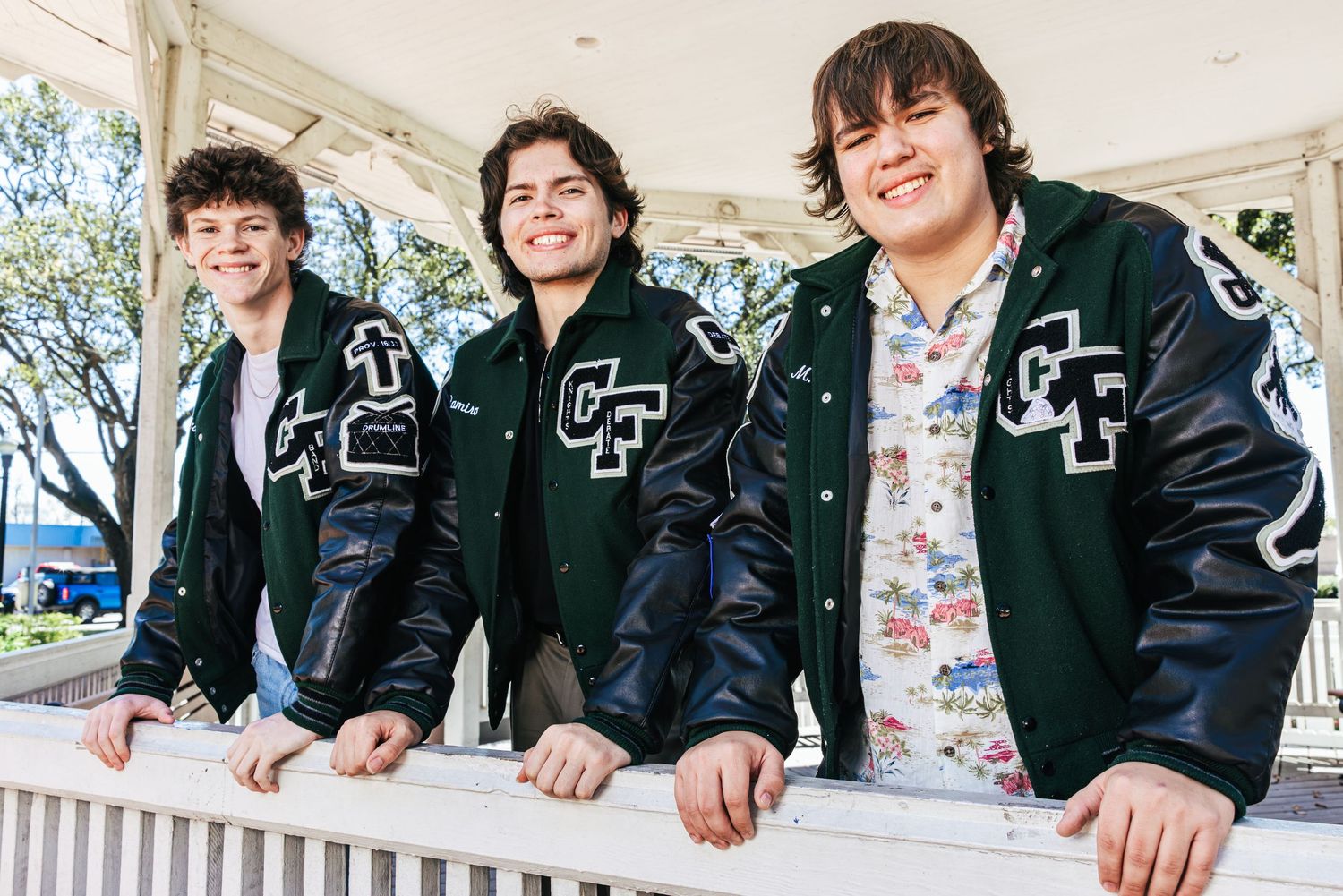 Three teenage boys wearing varsity jackets smile while leaning on a white railing outdoors.