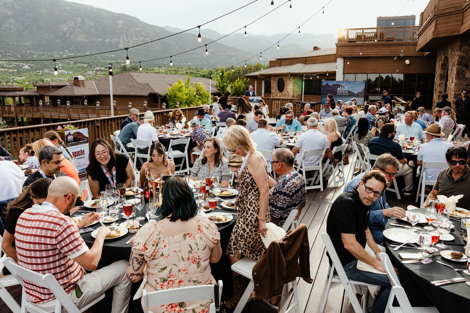Guests dining outdoors at a rooftop event with mountain views, string lights, and elegantly set tables.