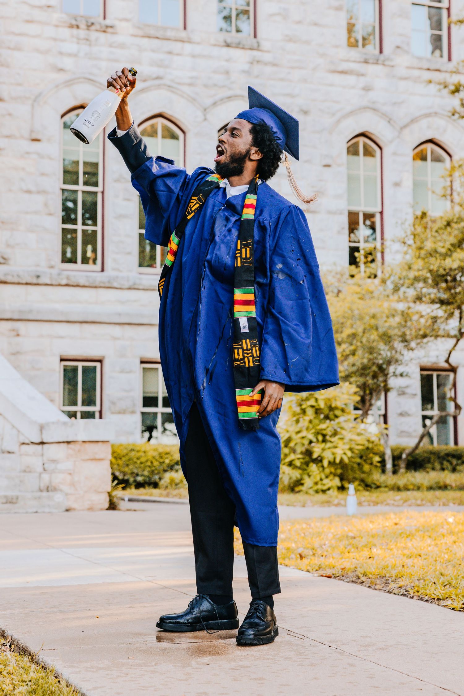 Graduate in blue cap and gown celebrates with joyful poses against stone building on sunny autumn day.