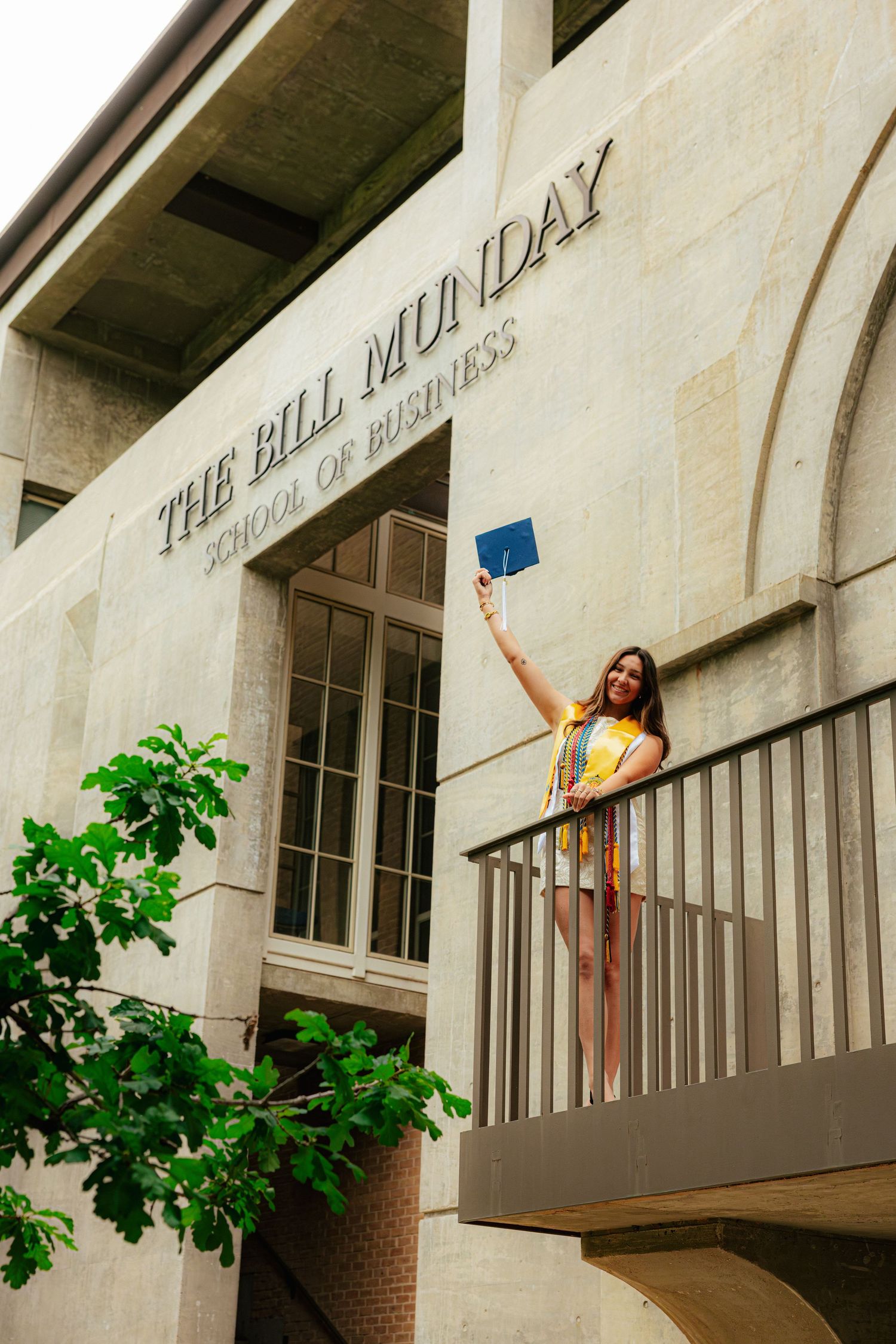 Graduate in yellow dress holds diploma aloft on balcony of The Bill Munday School of Business building.