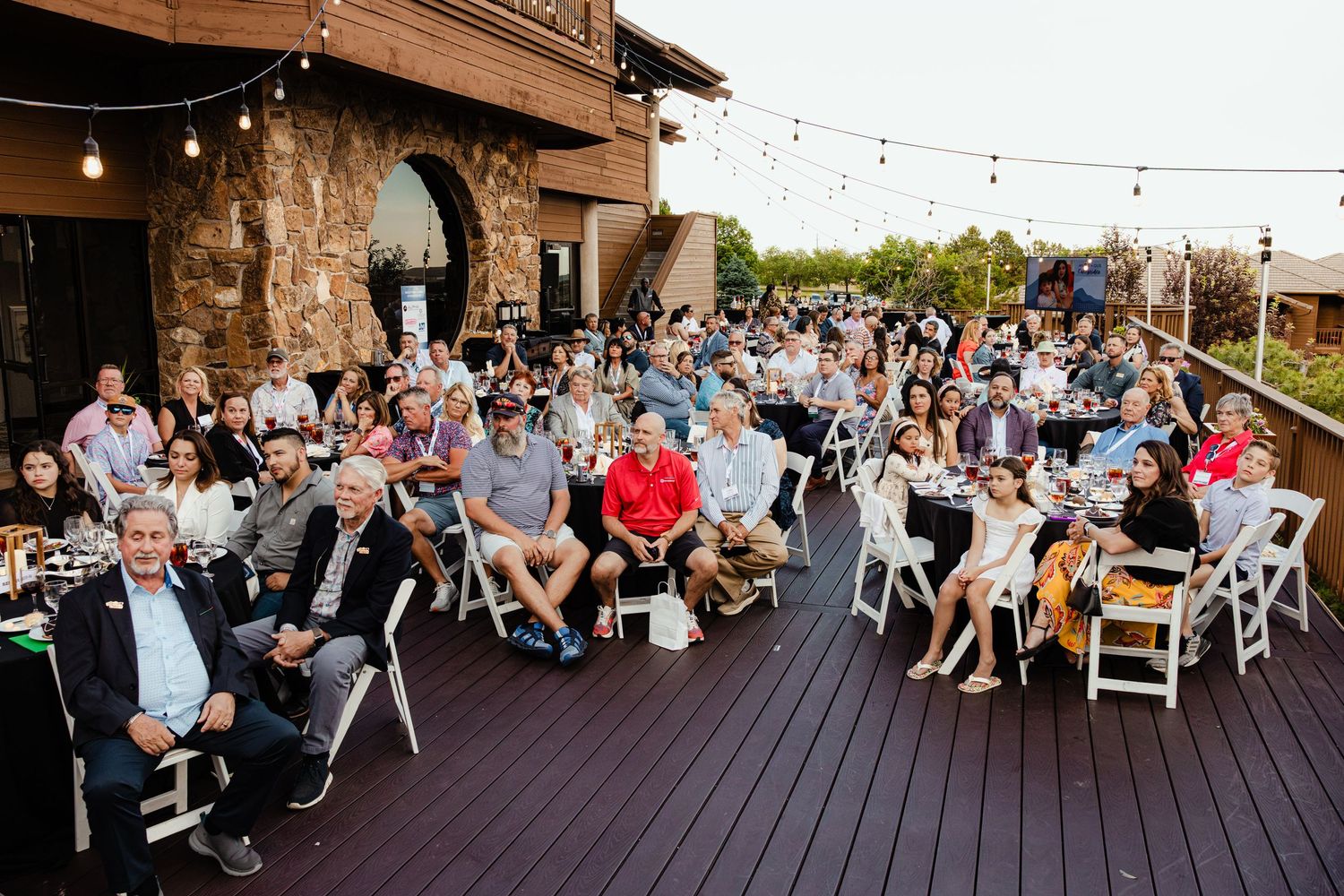 Large group of guests seated outdoors on a deck at an evening event, with string lights and a stone building backdrop.