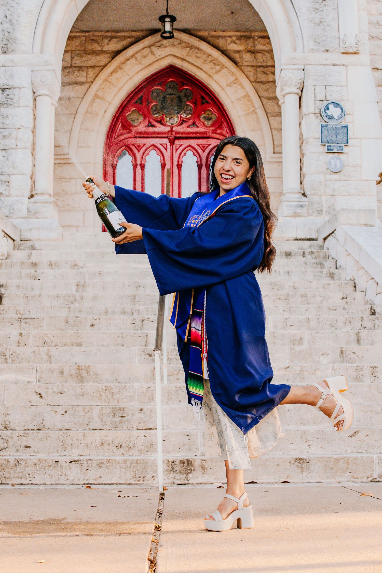 Graduate in blue academic regalia strikes celebratory dance poses in front of ornate stone archway.