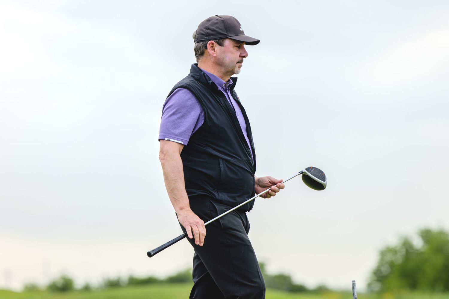 A golfer in a black vest follows through on their swing on a bright, sunny day at the golf course.