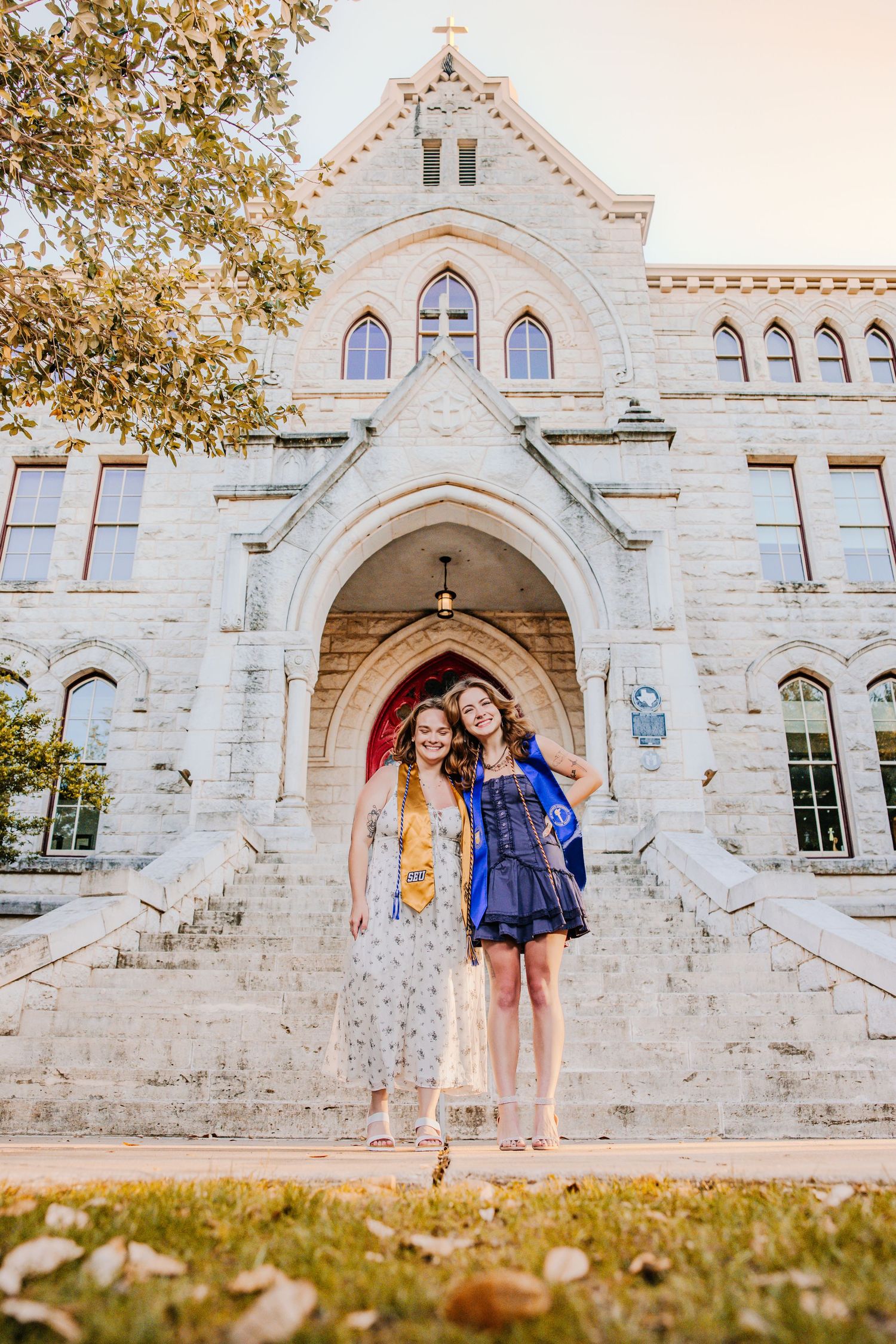 Two people pose together in front of a historic white stone church building with Gothic arched doorways at sunset.