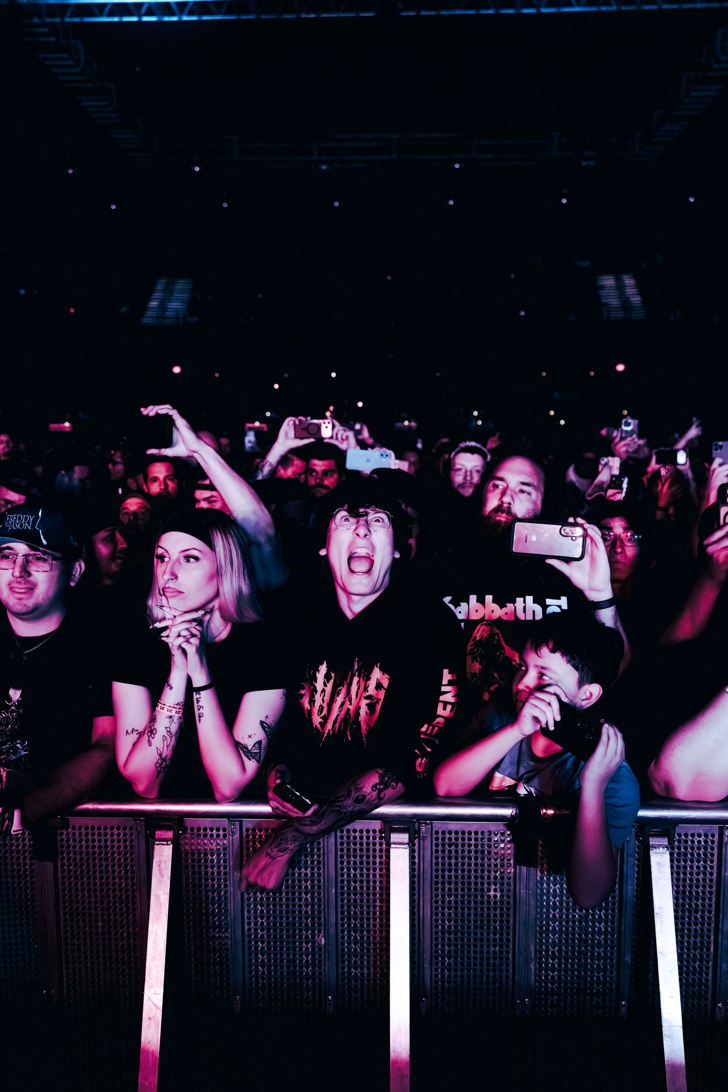 Excited concert crowd pressed against barrier at dark rock show, fans filming with phones under purple stage lighting.