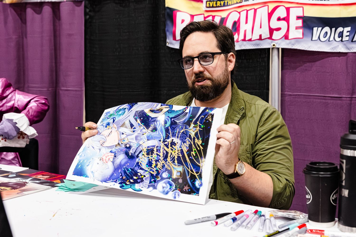 A bearded man in a green jacket holds colorful anime artwork at a comic convention booth.