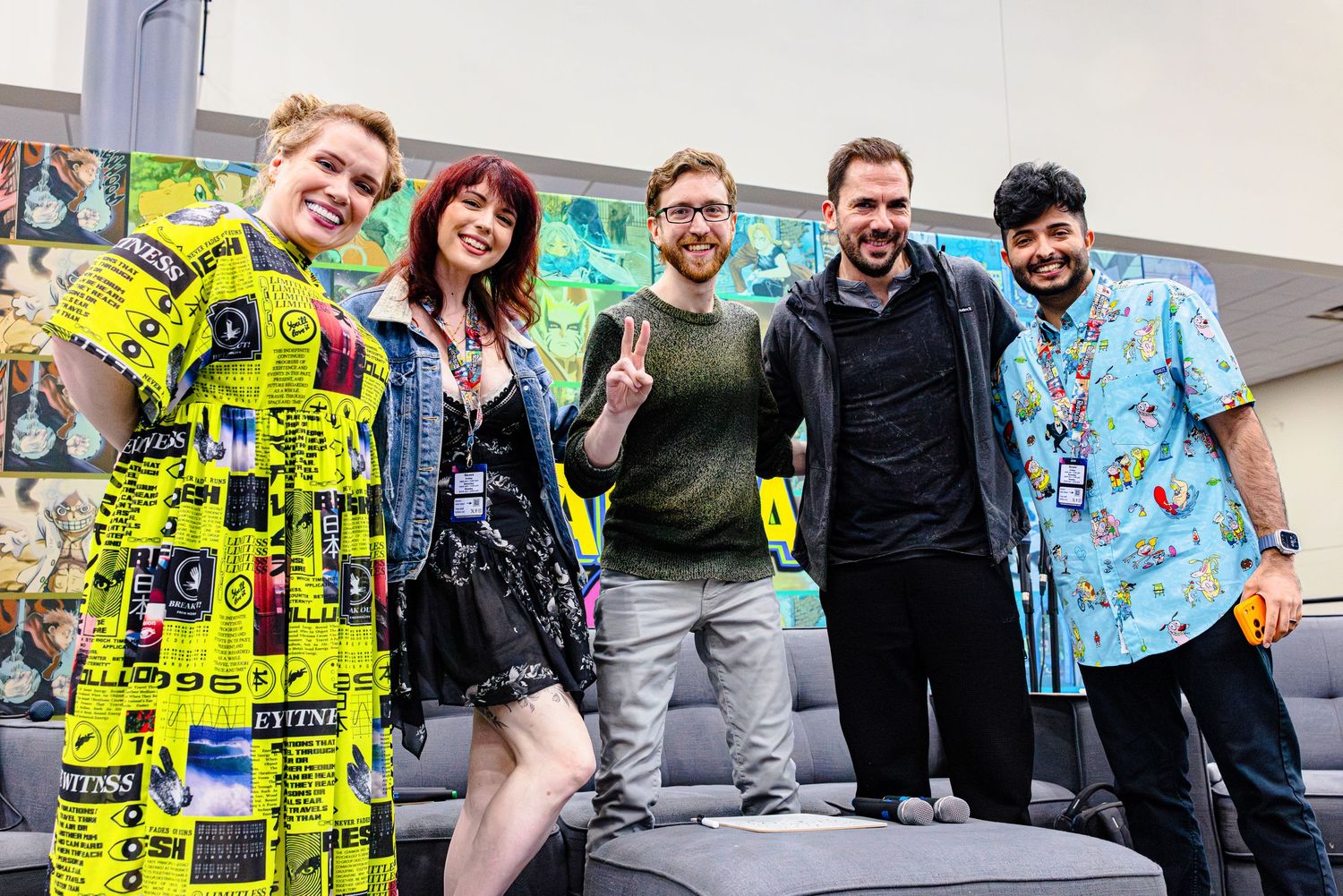 Five smiling people pose together at a colorful convention, wearing casual and vibrant outfits in a bright indoor setting.