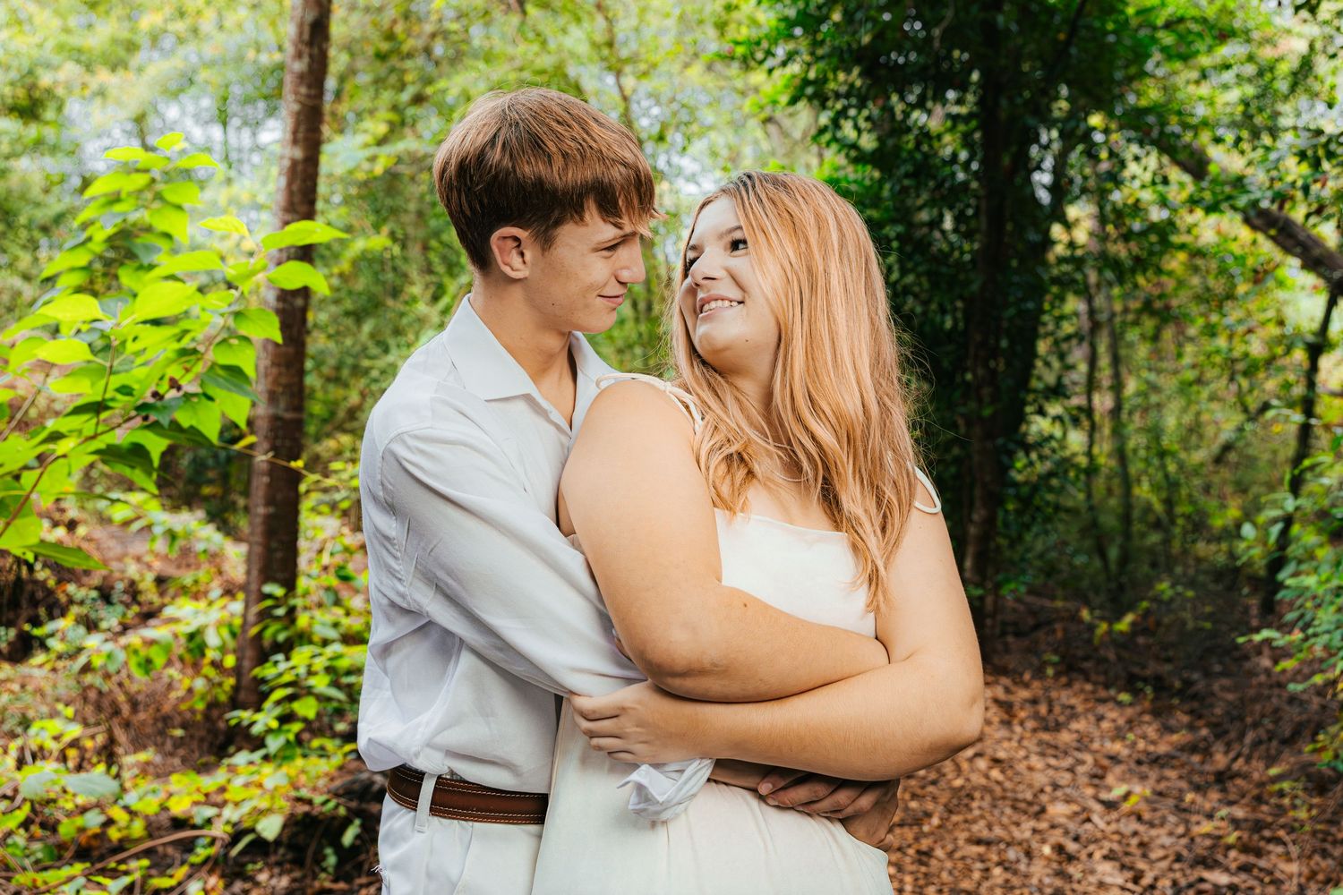 A couple in light colored outfits shares an intimate embrace while standing in a lush green wooded area.