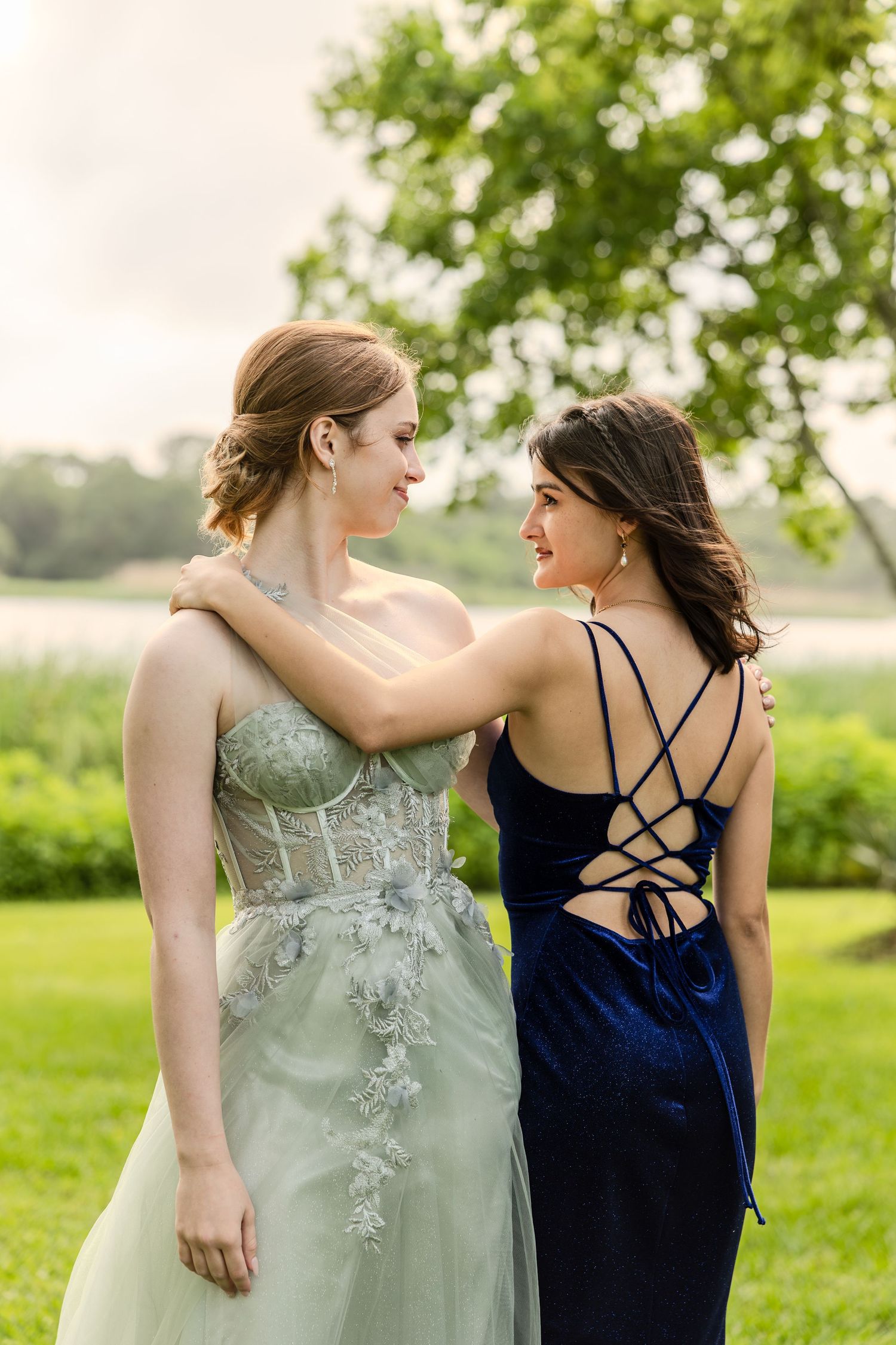 Two women in contrasting navy and sage formal dresses walk together in a lush green outdoor setting.