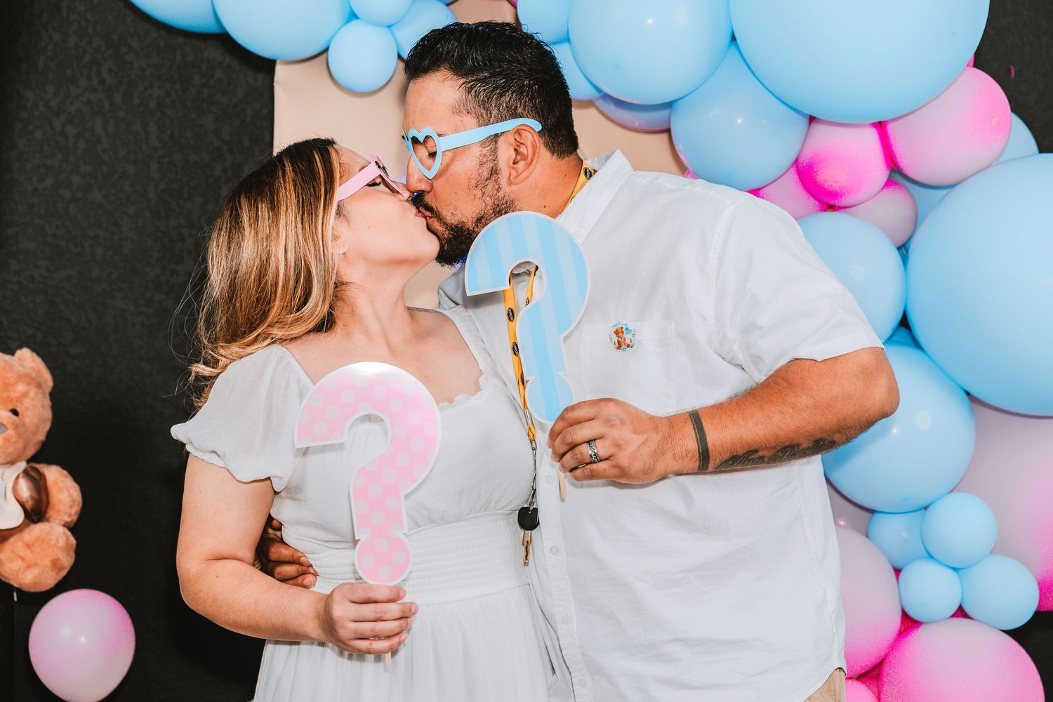 A couple shares a tender moment at a gender reveal party with pink and blue balloon decorations in the background.