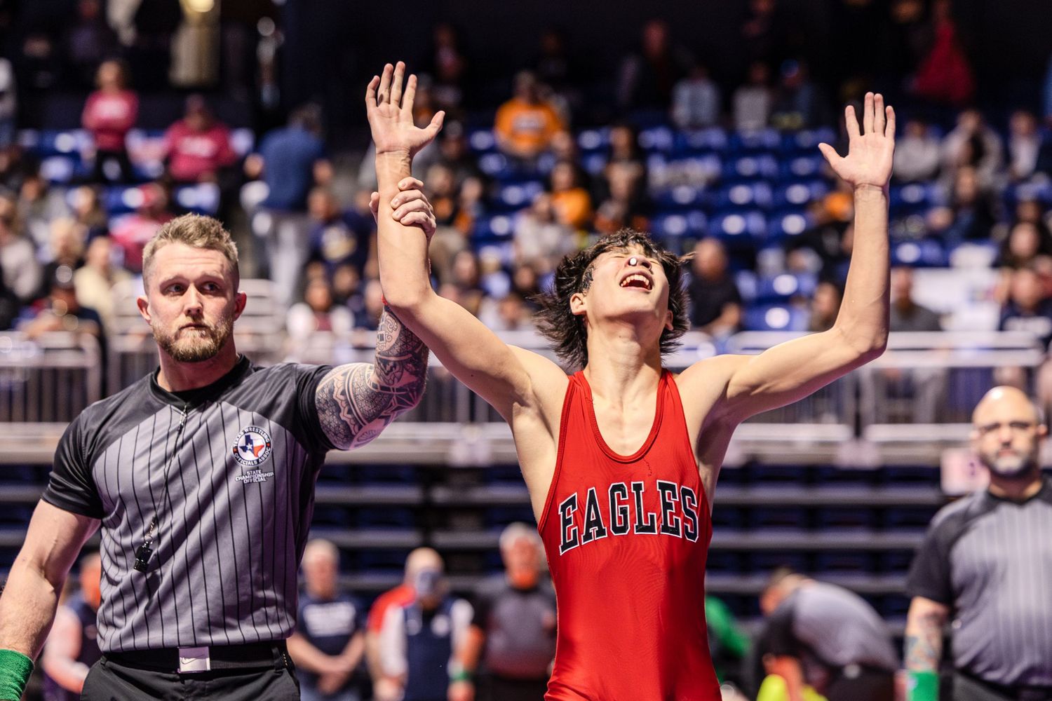 A wrestler in a red singlet celebrates victory with arms raised while a referee signals the win in an arena setting.